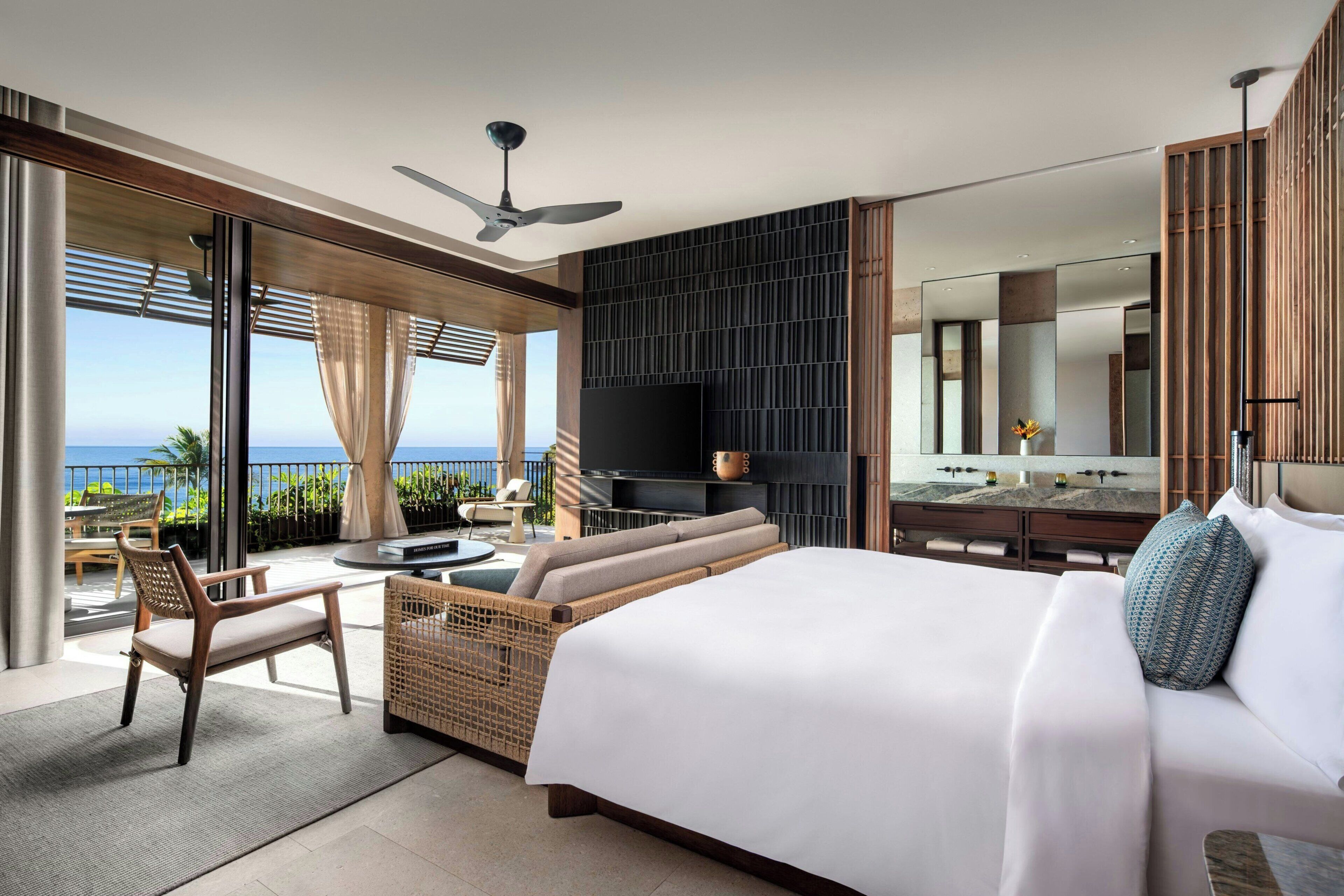 A modern hotel room with a large bed, a seating area with a view of the ocean, a flat-screen TV, and a balcony with outdoor furniture.