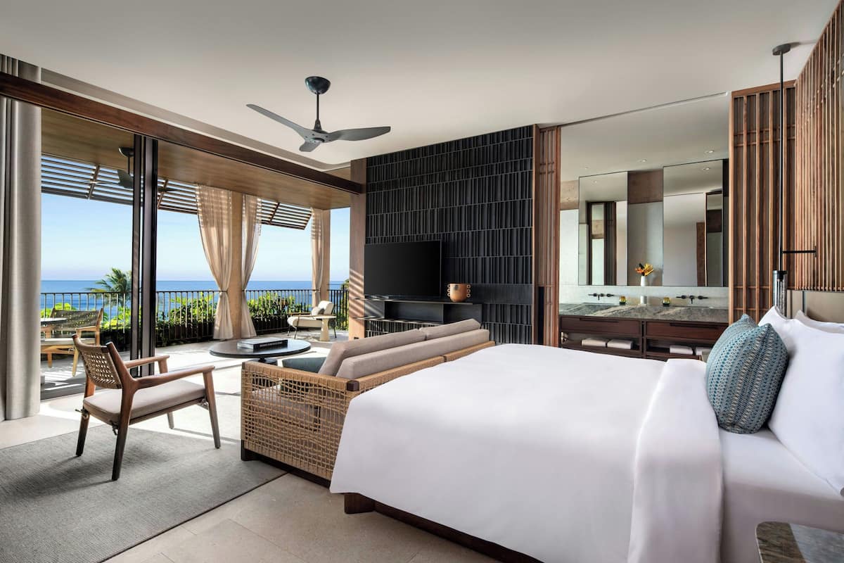 Une chambre d’hôtel moderne dotée d’un grand lit, d’un coin salon avec vue sur l’océan, d’une télévision à écran plat et d’un balcon aménagé avec du mobilier d’extérieur.