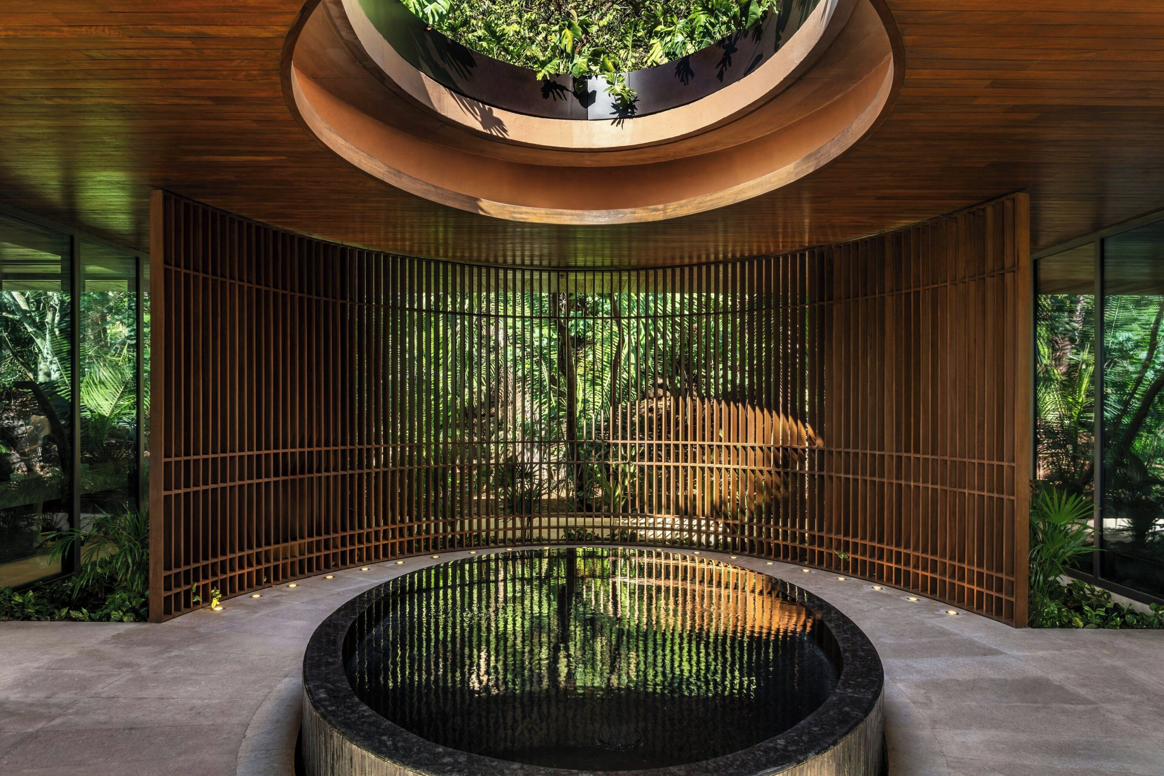 Un elemento acuático circular con techo de madera y vista a un bosque a través de una celosía de bambú.
