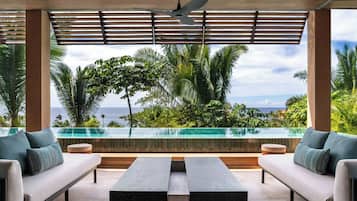 Un área de estar moderna al aire libre con vista a la piscina, un ventilador de techo y un exuberante jardín tropical de fondo.