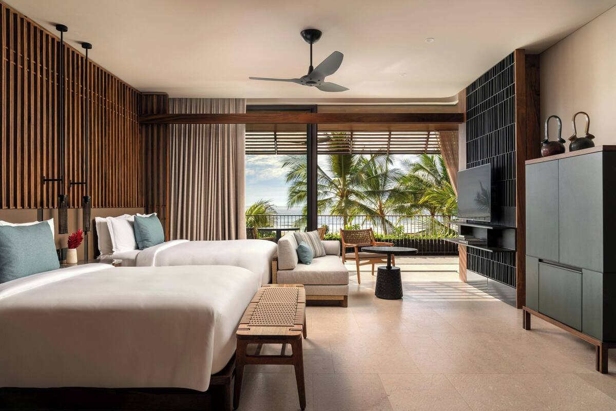 A hotel room with two beds, a ceiling fan, a TV, a balcony with a view of palm trees, and a large cabinet.