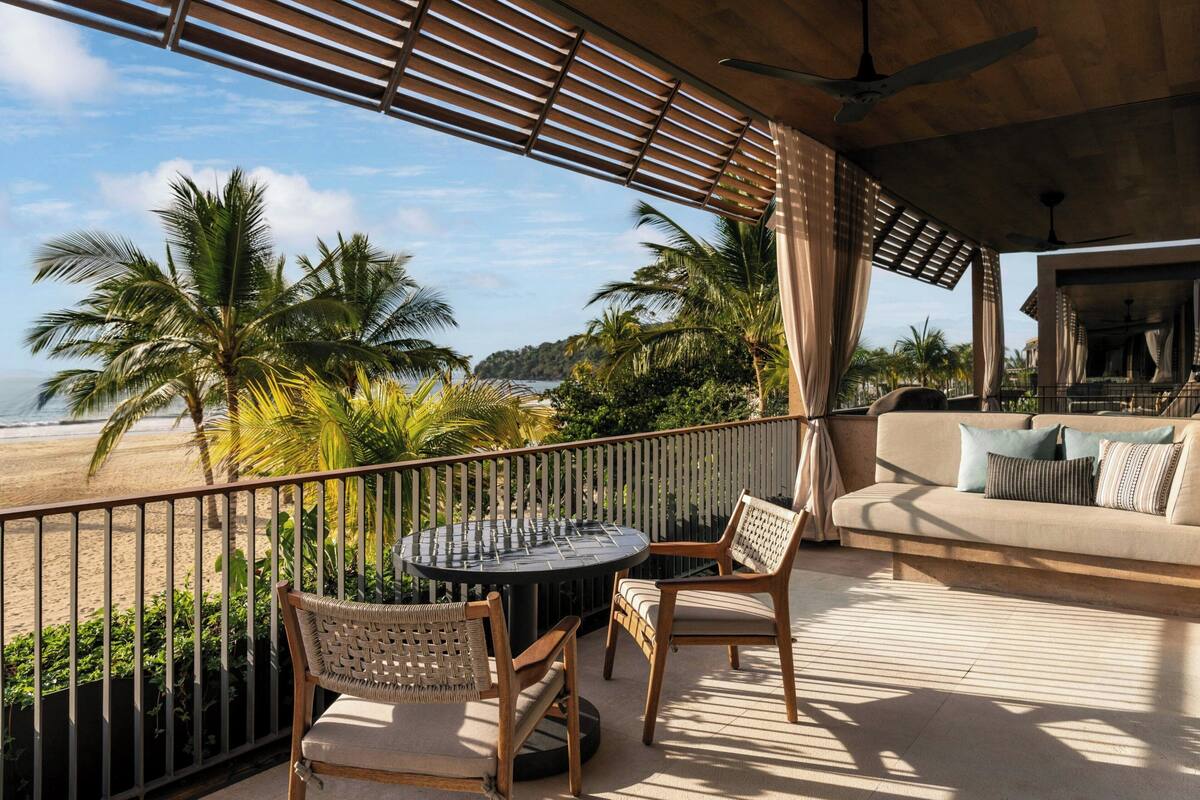 A balcony with a view of the beach, a round table, two wicker chairs, and a sofa with cushions.
