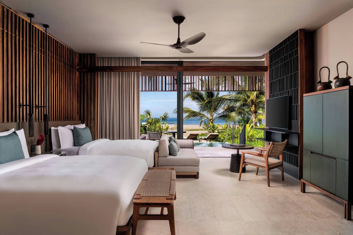 A hotel room with two beds, a sofa, a TV, and a view of the beach.