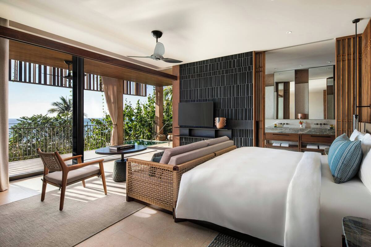 A modern hotel room with a large bed, a sofa, a television, and a dining area with a view of the outdoors.