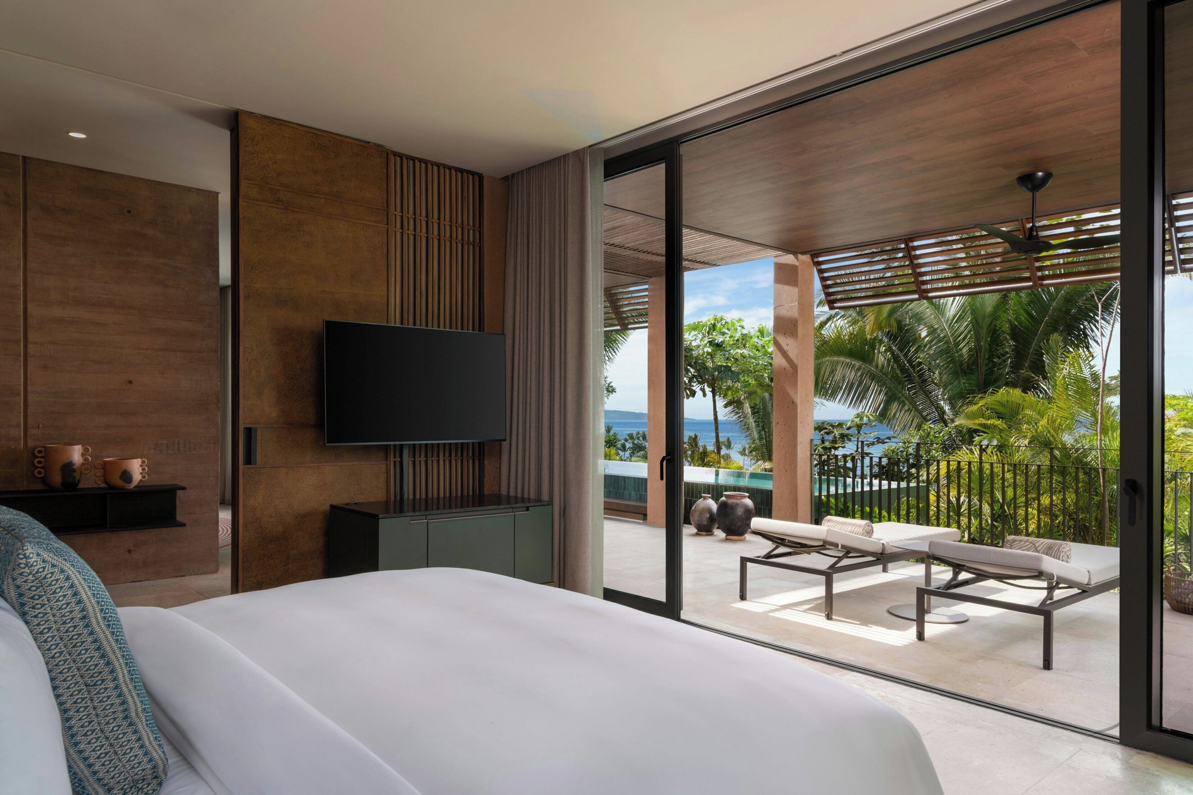A modern hotel room with a large bed, a flat-screen TV, a balcony with outdoor seating, and a view of the ocean.
