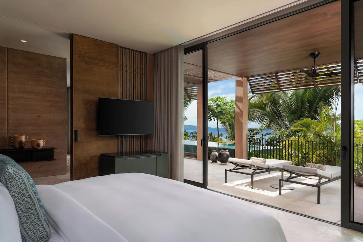 A modern hotel room with a large bed, a flat-screen TV, a balcony with outdoor seating, and a view of the ocean.