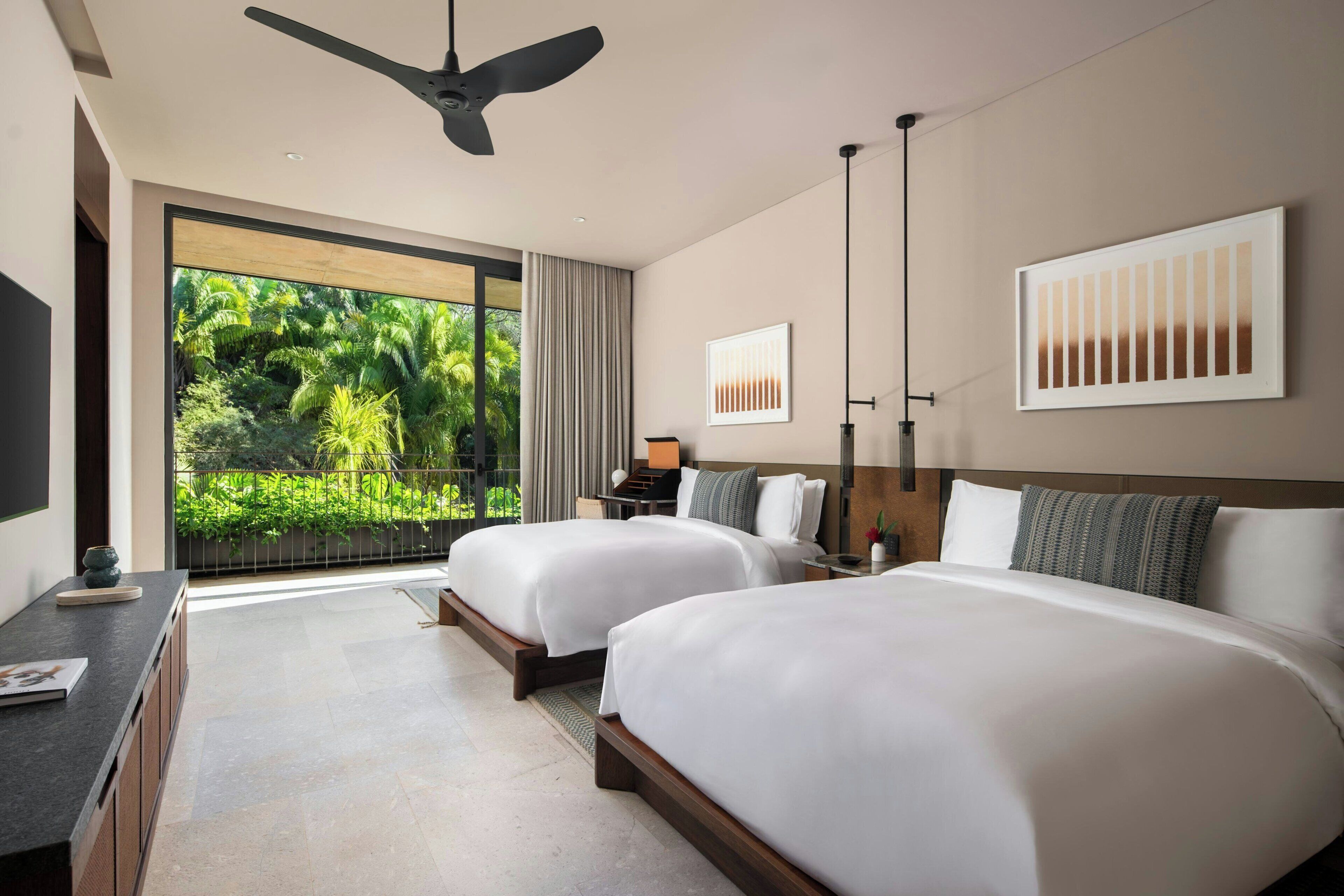 Habitación de hotel con dos camas, ventilador de techo, ventana grande con vistas a zonas verdes y obras de arte en las paredes.