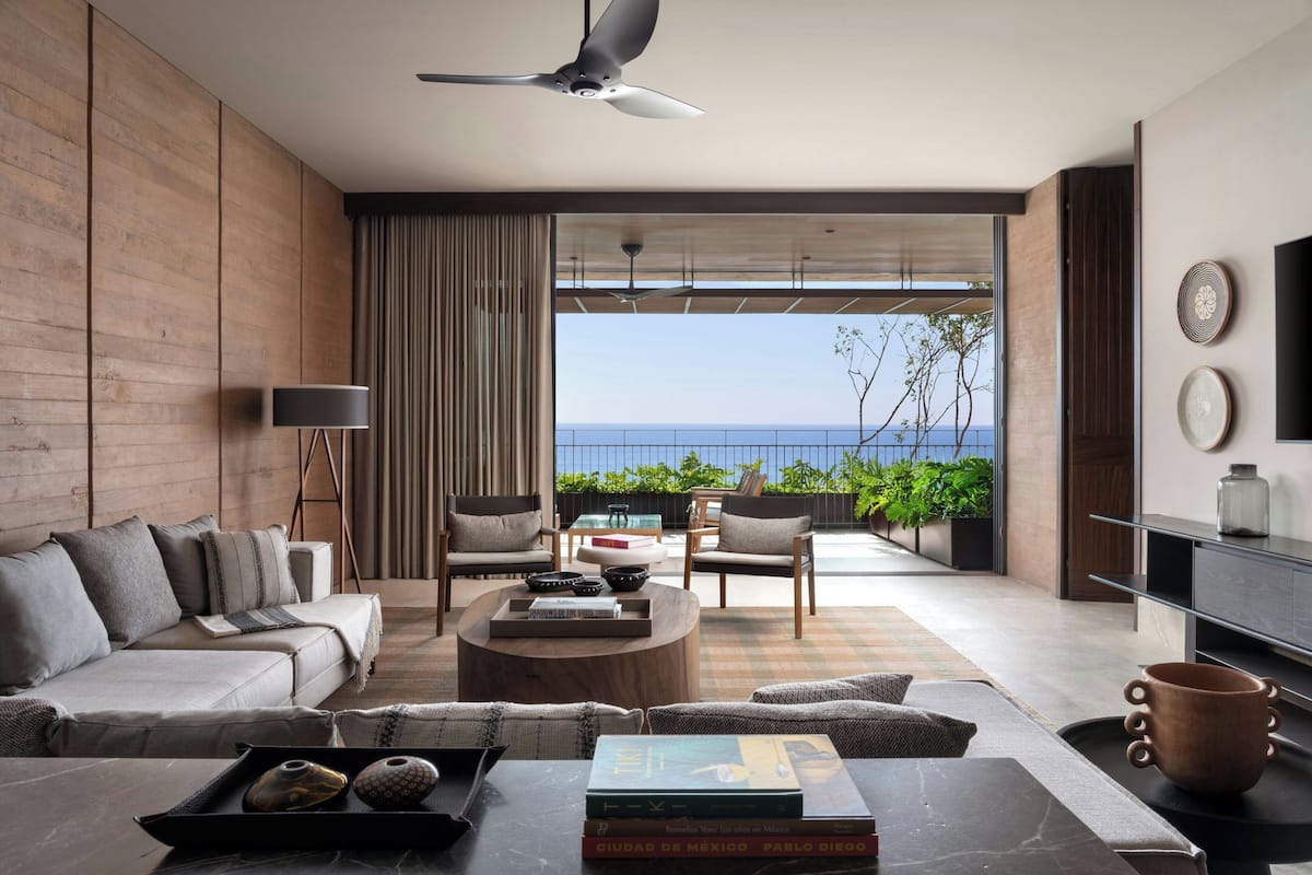 A modern living room with a sofa, coffee table, and chairs, a ceiling fan, and a view of the sea.