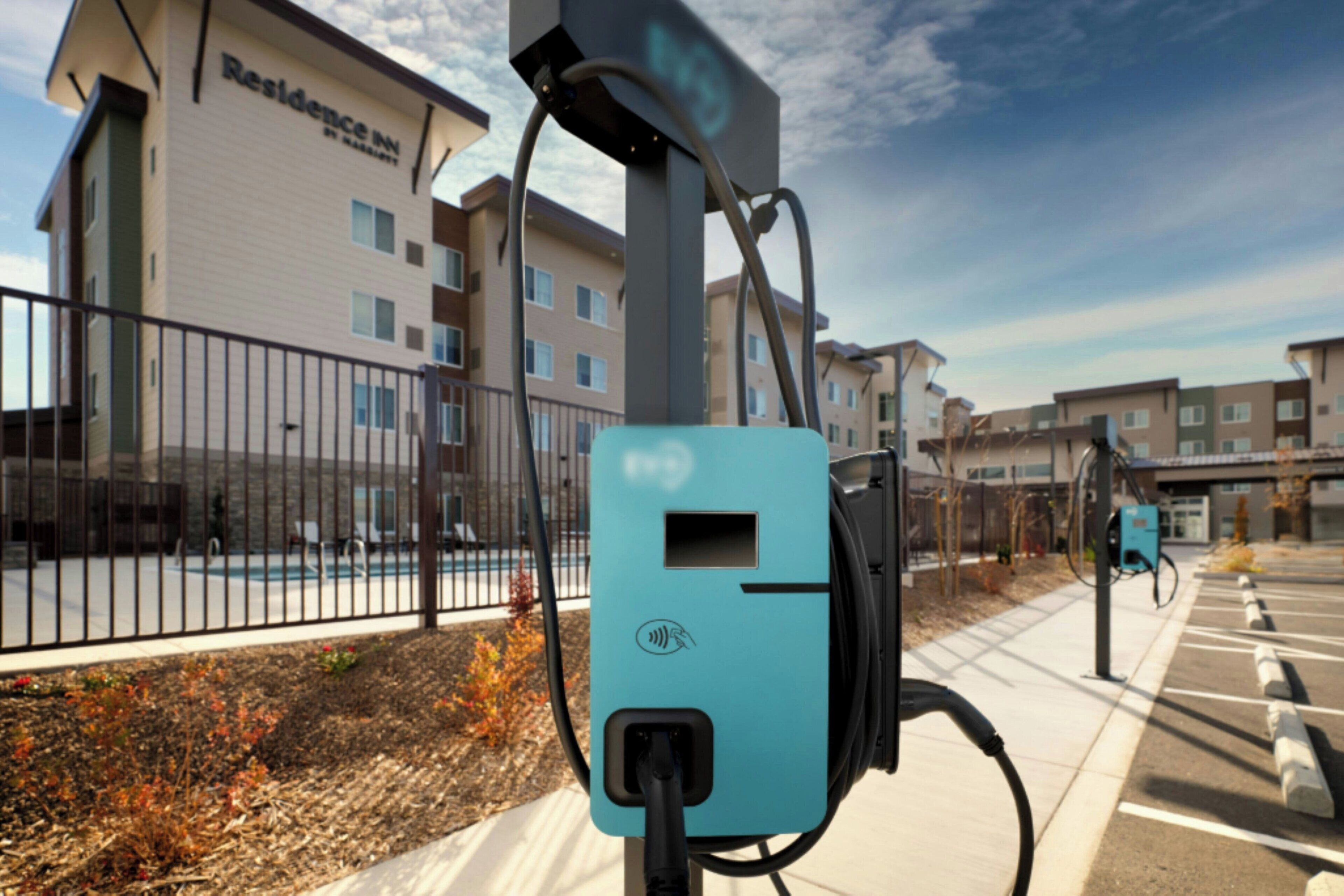 A parking area with electric vehicle charging stations, a fence, and residential buildings in the background.