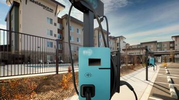 A parking area with electric vehicle charging stations, a fence, and residential buildings in the background.