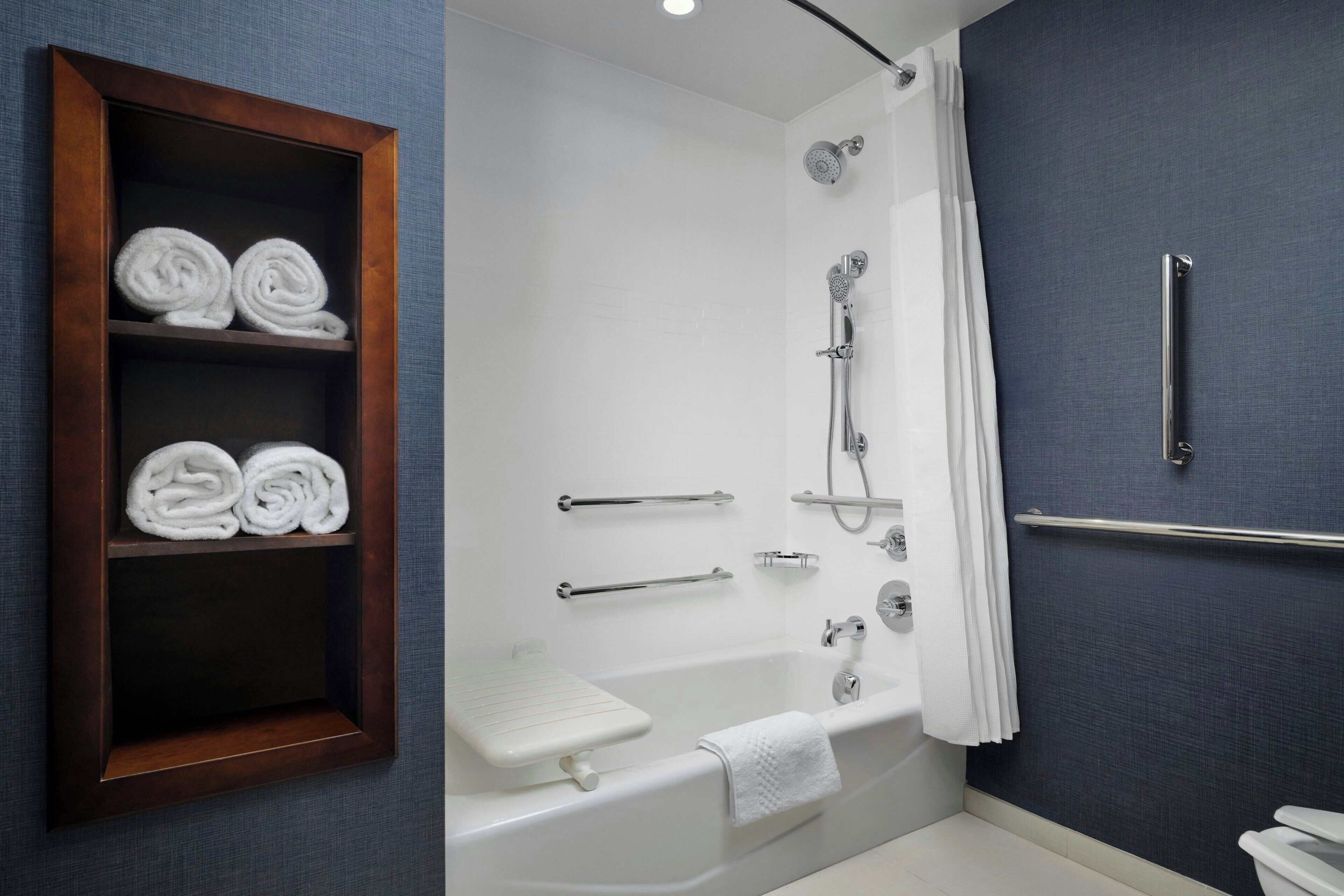A modern bathroom with a bathtub, shower, and toilet. There are towels neatly rolled and placed in a wooden shelf.