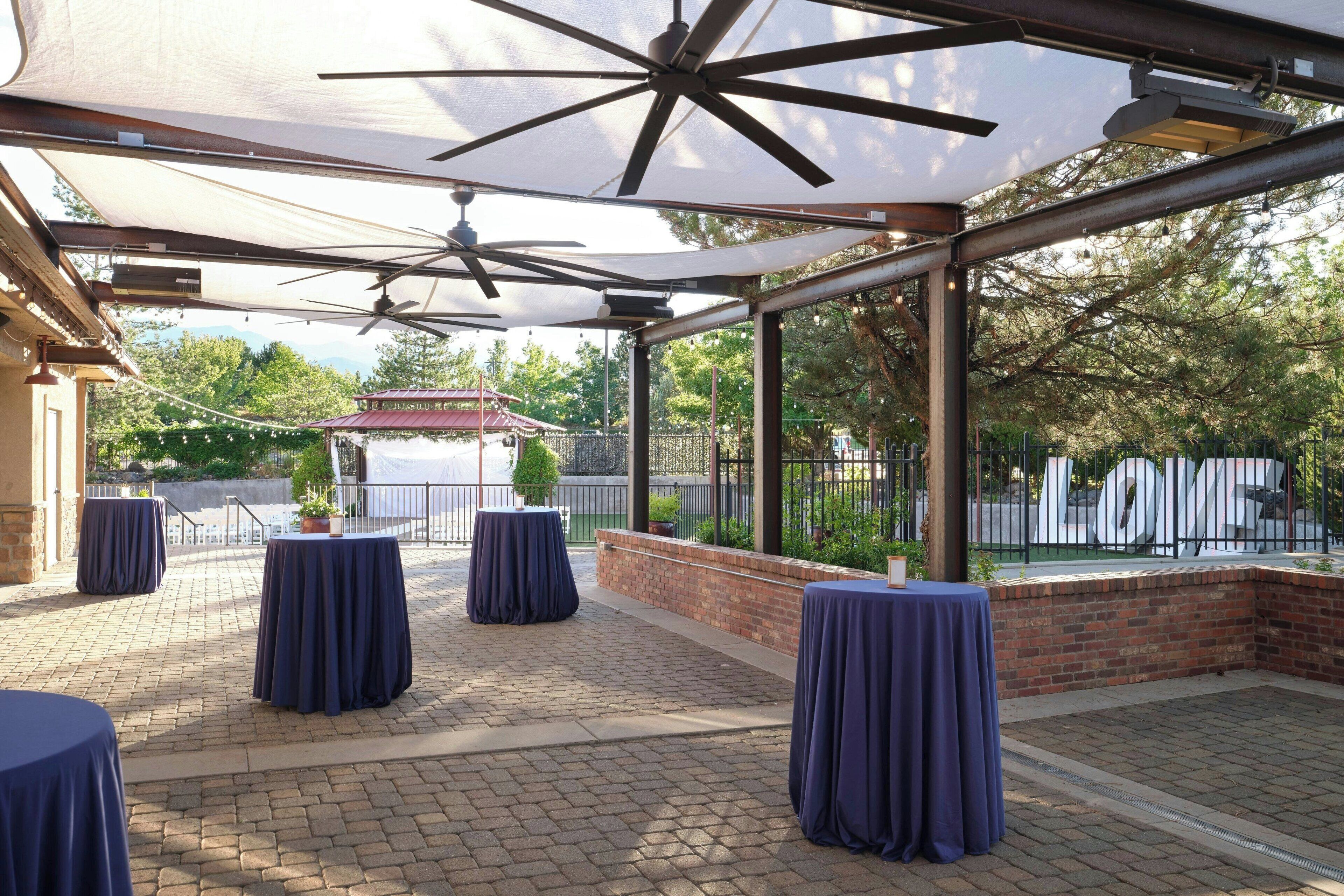 An outdoor patio area with brick flooring, covered by a canopy with ceiling fans, and tables with dark tablecloths.