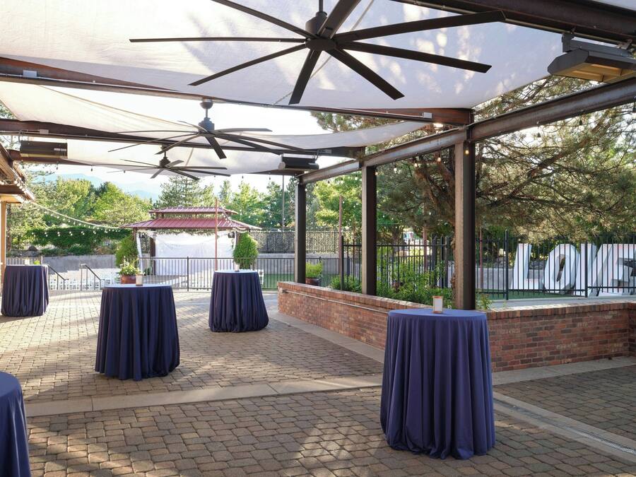 An outdoor patio area with brick flooring, covered by a canopy with ceiling fans, and tables with dark tablecloths.