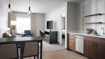 A modern hotel room with a dining area, a TV, a flat-screen TV mounted on the wall, a kitchen area with a dishwasher, and a large window with curtains.