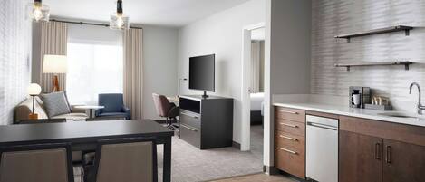 A modern hotel room with a dining area, a TV, a flat-screen TV mounted on the wall, a kitchen area with a dishwasher, and a large window with curtains.