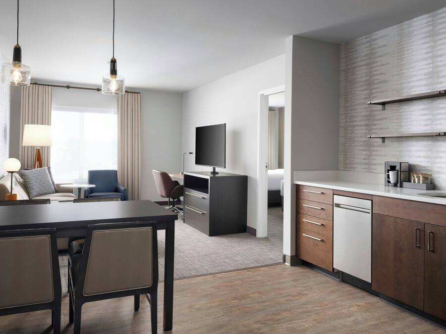 A modern hotel room with a dining area, a TV, a flat-screen TV mounted on the wall, a kitchen area with a dishwasher, and a large window with curtains.