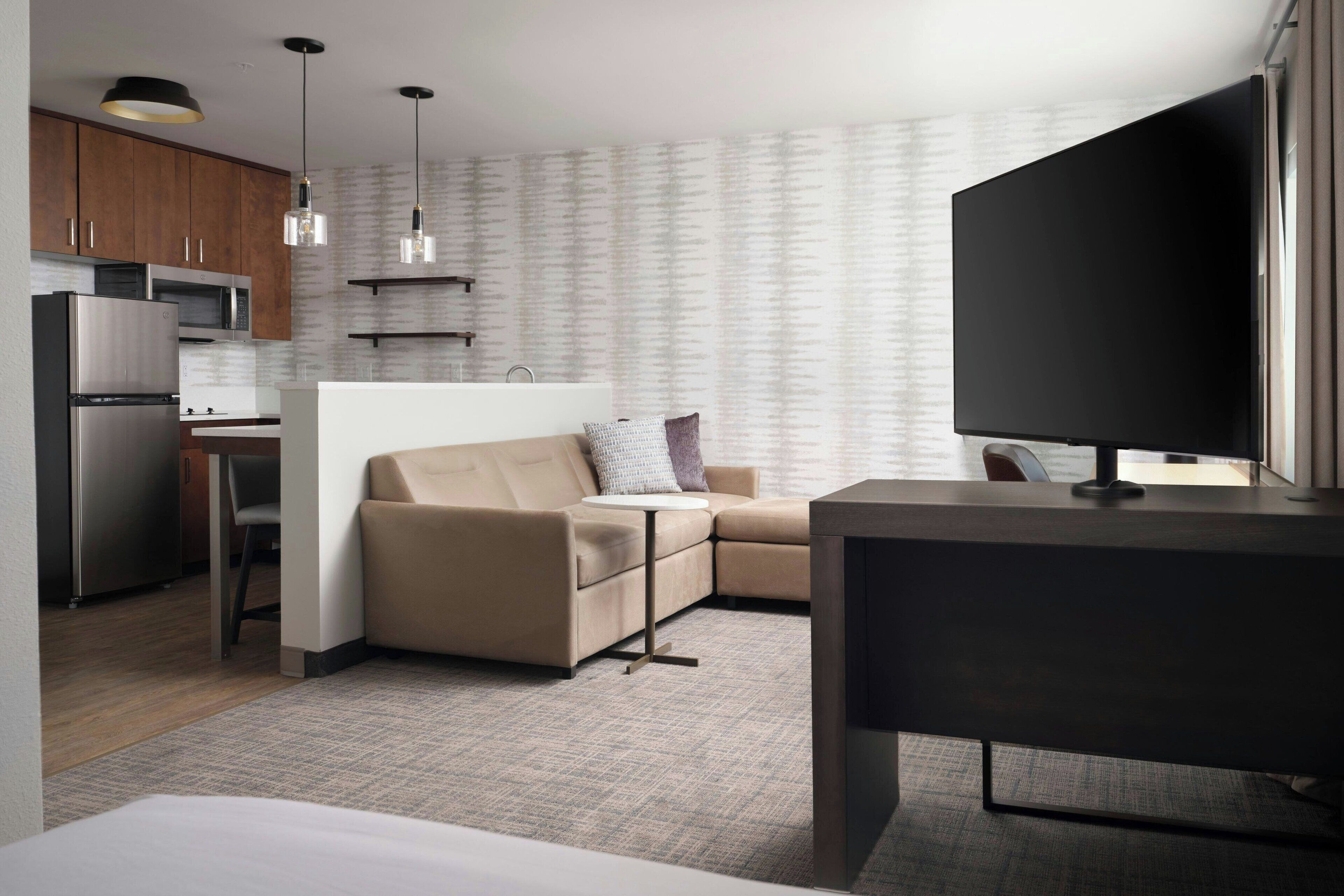 A modern hotel room with a kitchenette, a sofa, a TV, and a desk.