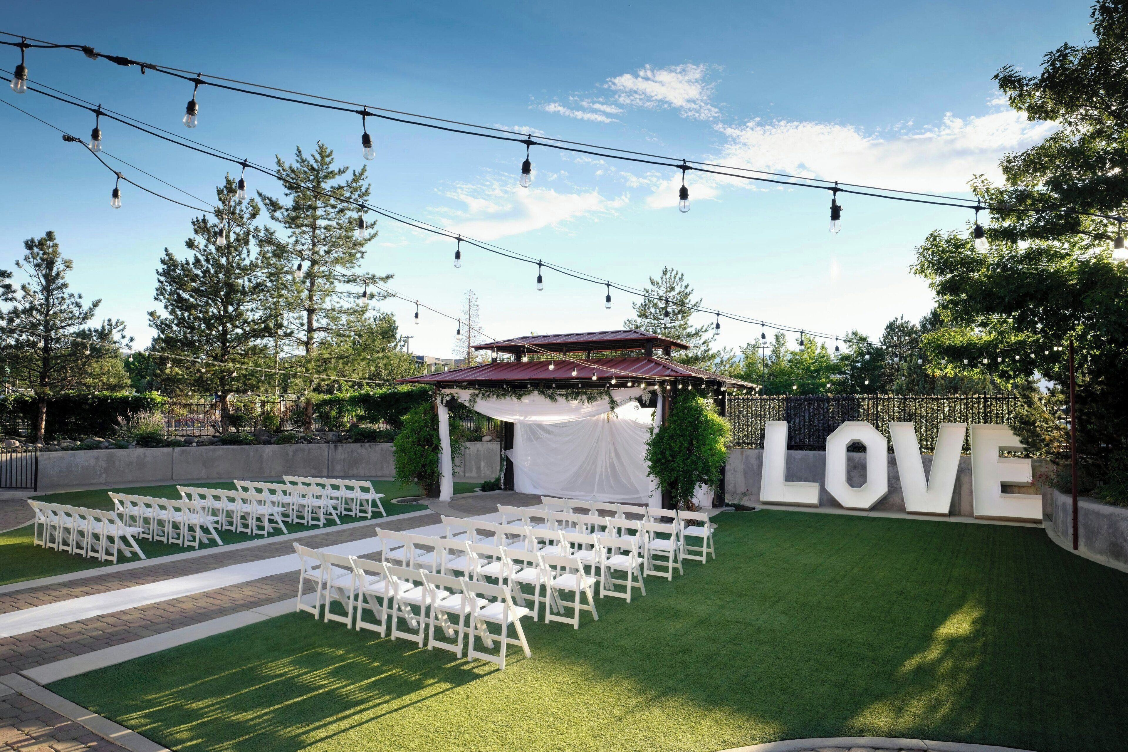 An outdoor wedding venue with white chairs arranged in rows, a stage with draped white fabric, and a large 'LOVE' sign.