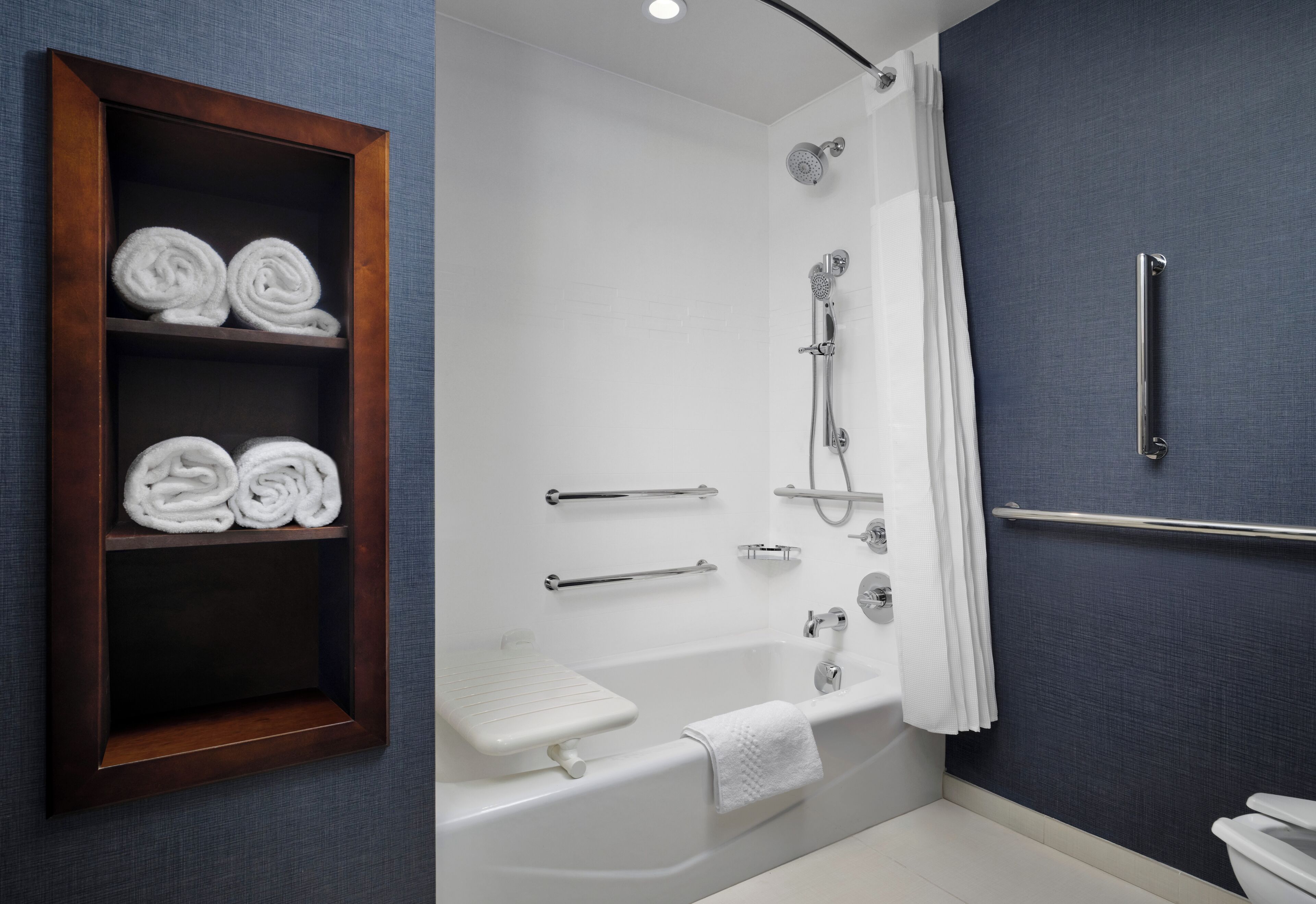 Een moderne badkamer met een bad, douche en toilet. Handdoeken liggen op een plank en er zit een handgreep aan de muur.