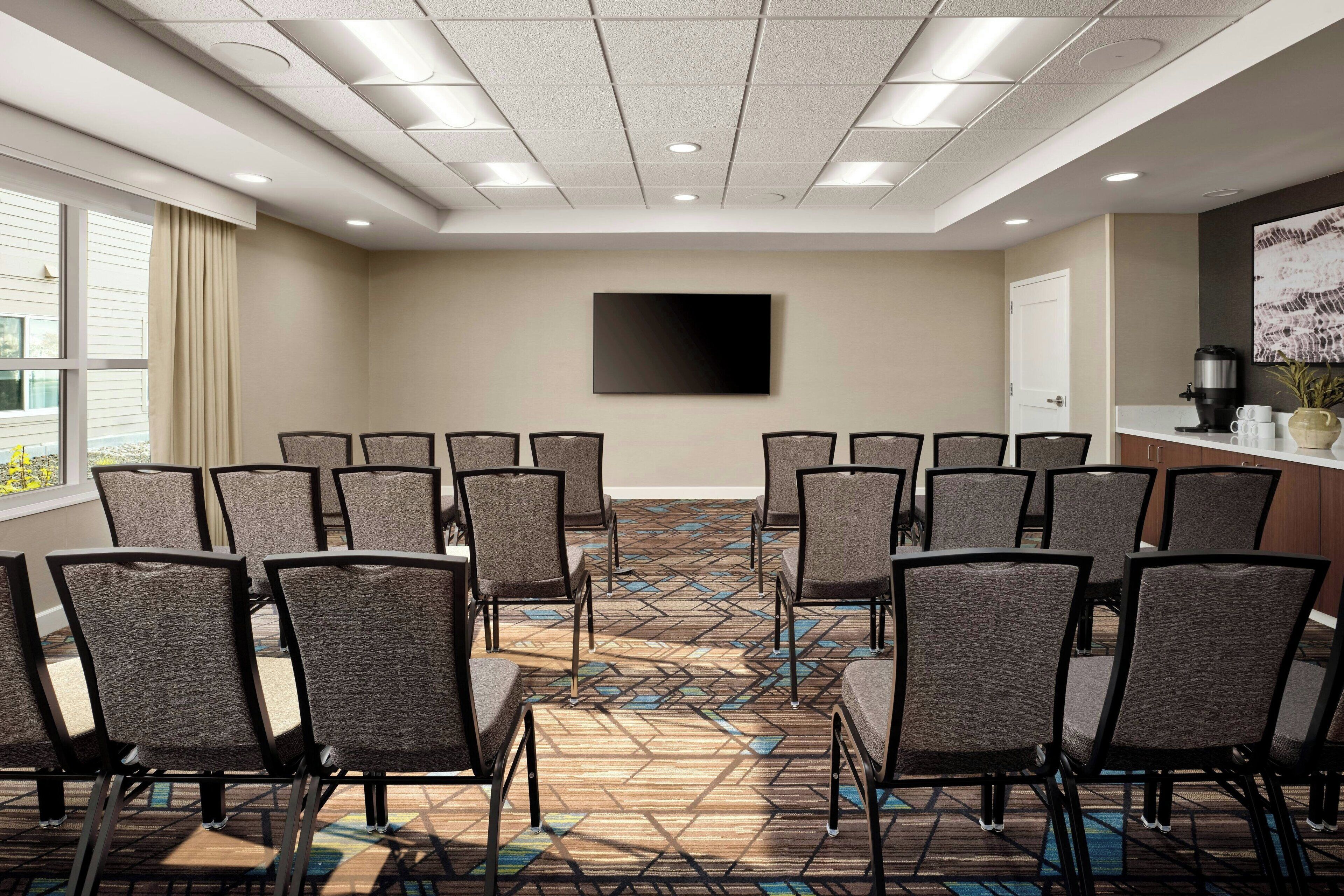 A conference room with 12 chairs arranged in a semi-circle, a large flat-screen TV on the wall, and a coffee machine on a counter.