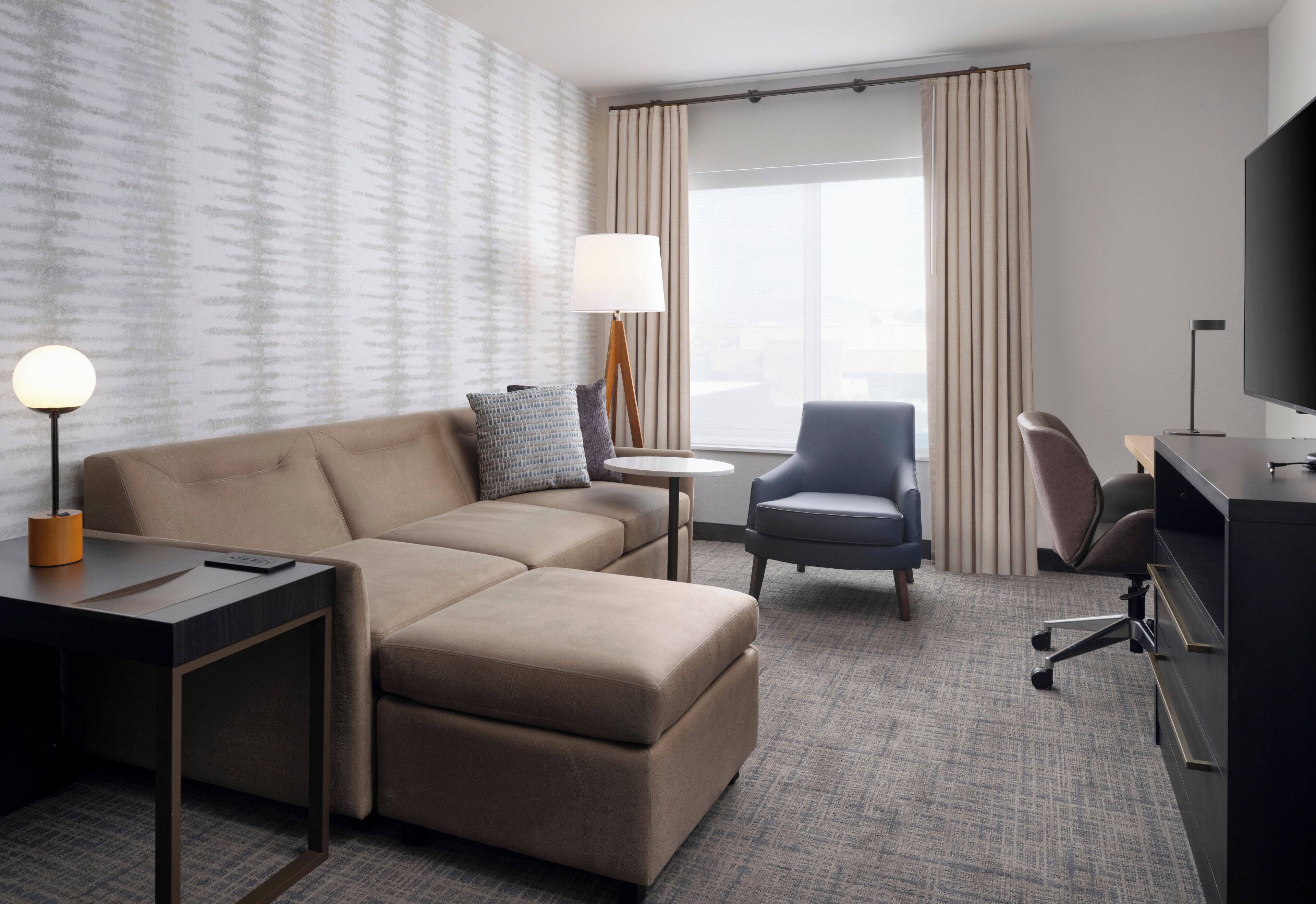A hotel room with a sofa, armchair, desk, lamp, TV, and a window with curtains.