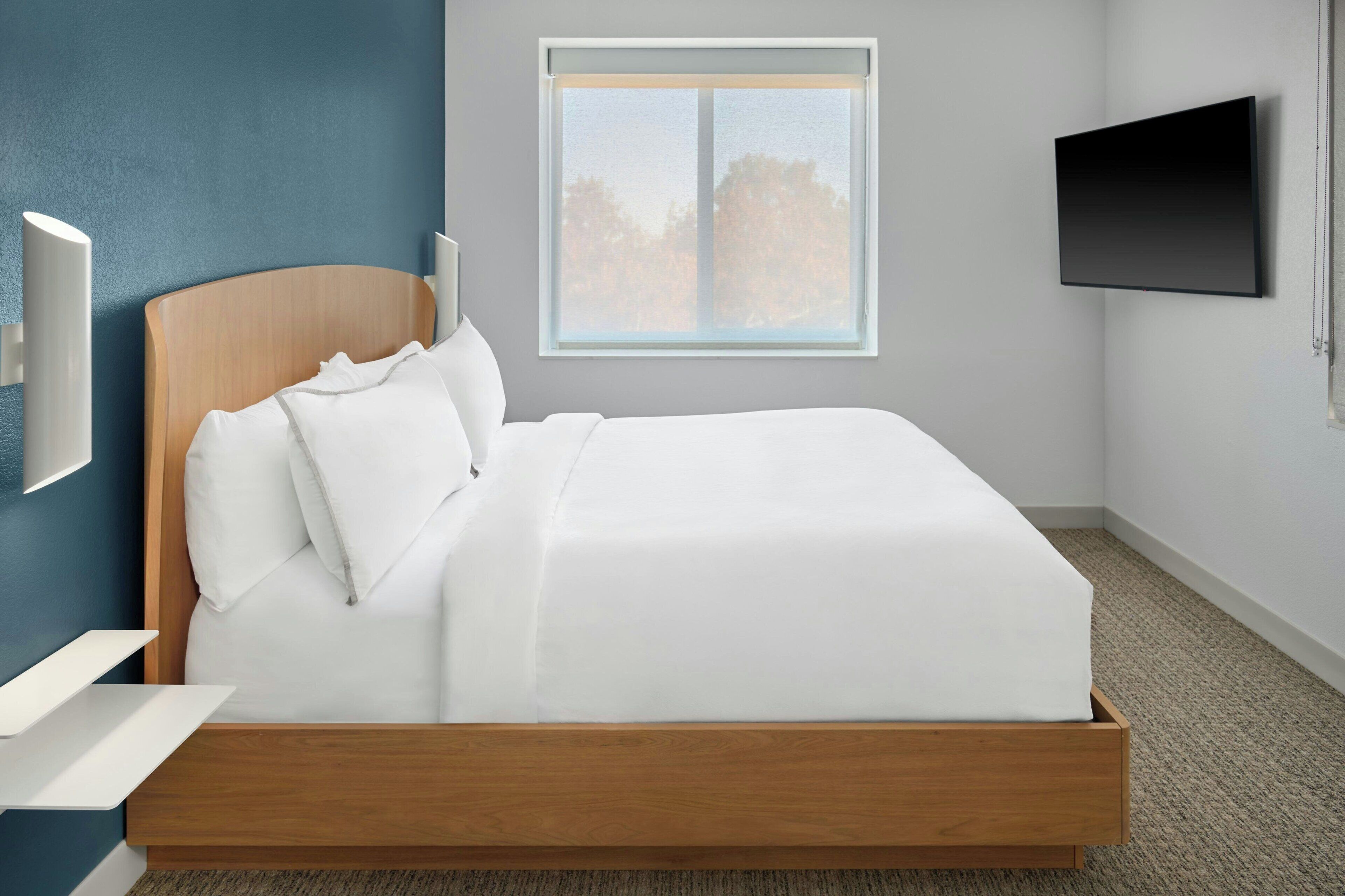 A modern bedroom with a wooden bed frame, white bedding, a mounted TV, and a window with a view of trees.