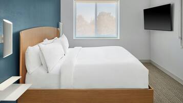 A modern bedroom with a wooden bed frame, white bedding, a mounted TV, and a window with a view of trees.