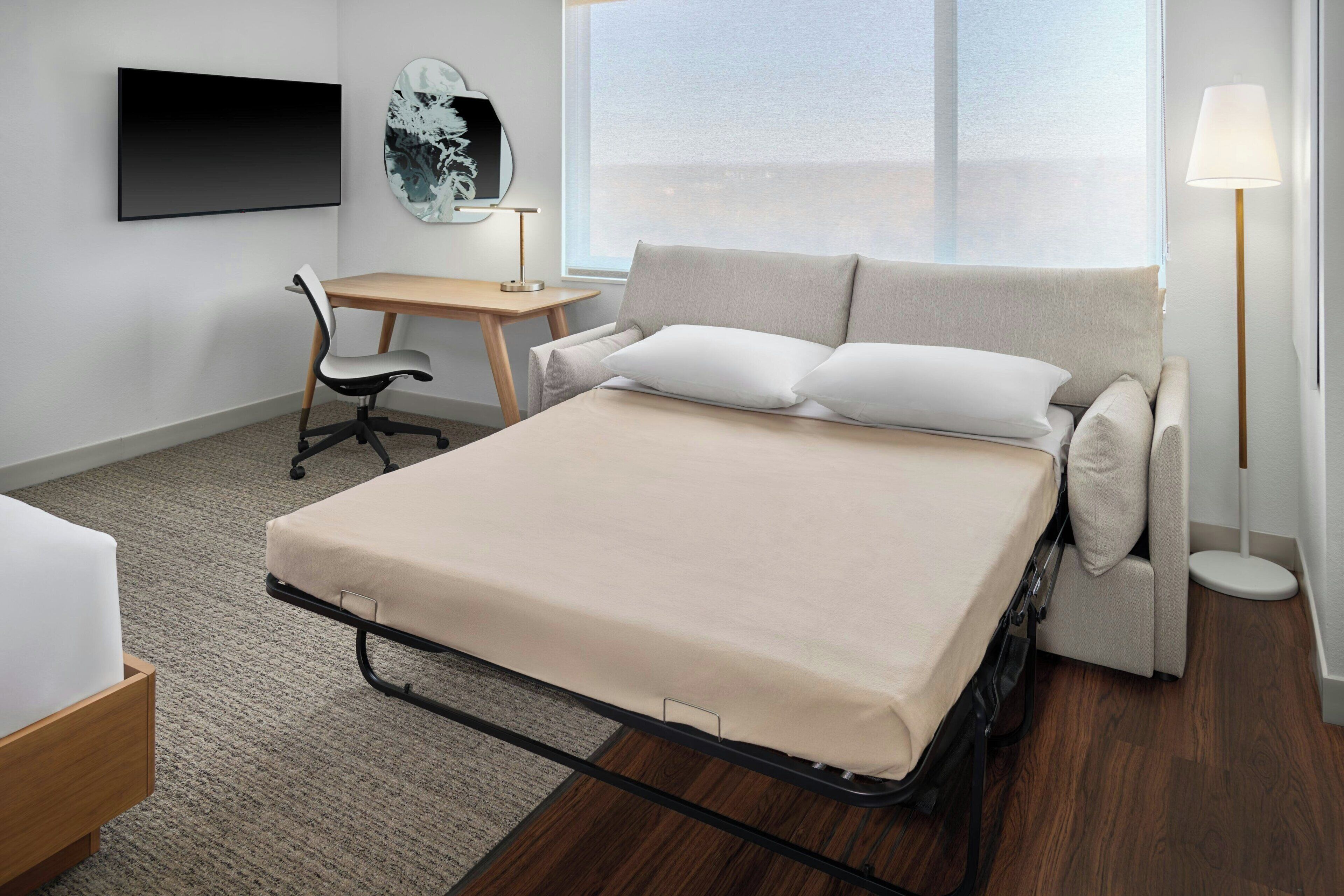 A hotel room with a bed, a desk, a chair, a lamp, and a large window.
