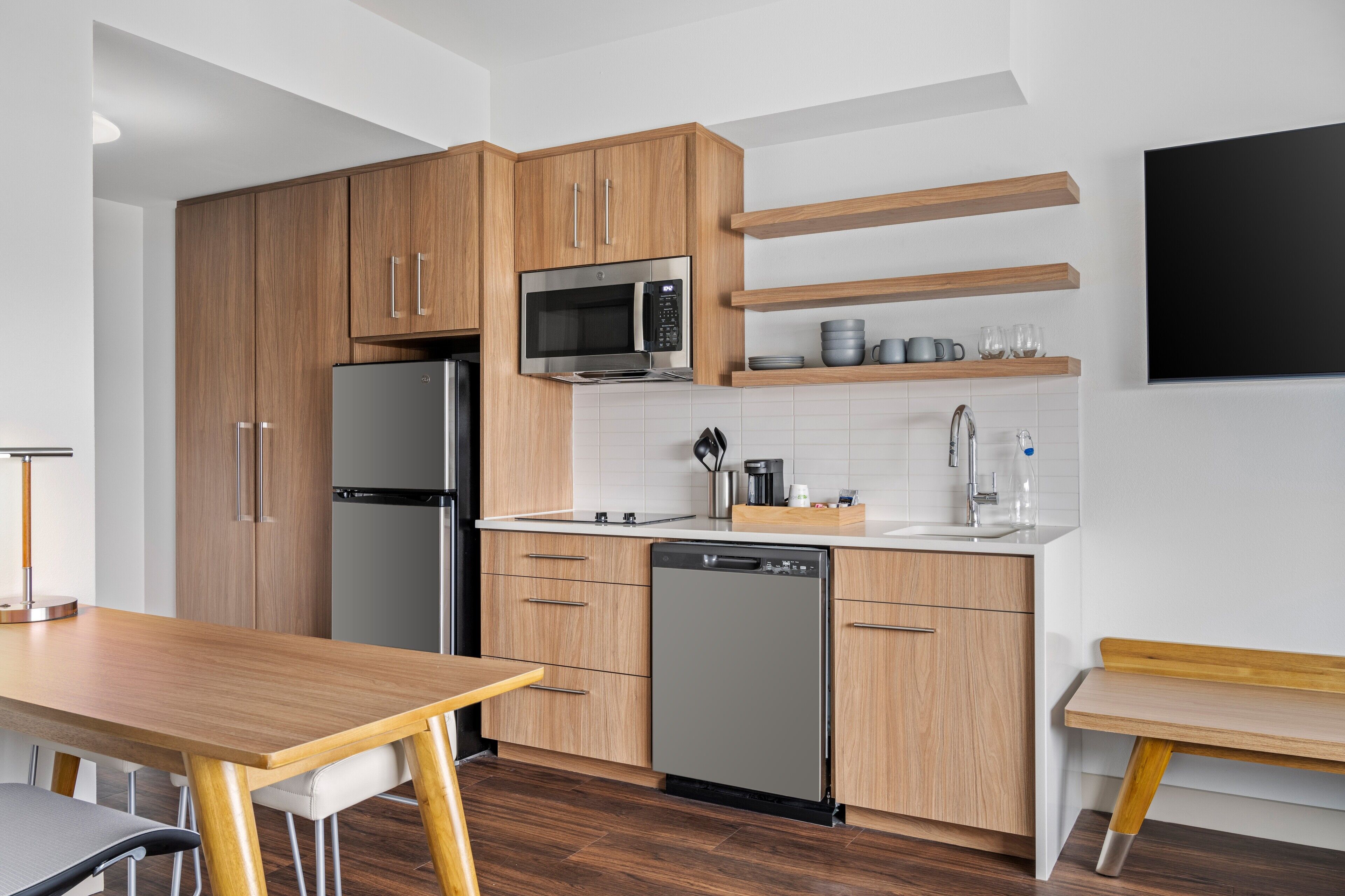 Une cuisine moderne avec des meubles en bois, un réfrigérateur en acier inoxydable, un four à micro-ondes et un lave-vaisselle. On trouve des étagères en bois au-dessus de l’évier et un téléviseur à écran plat fixé au mur.