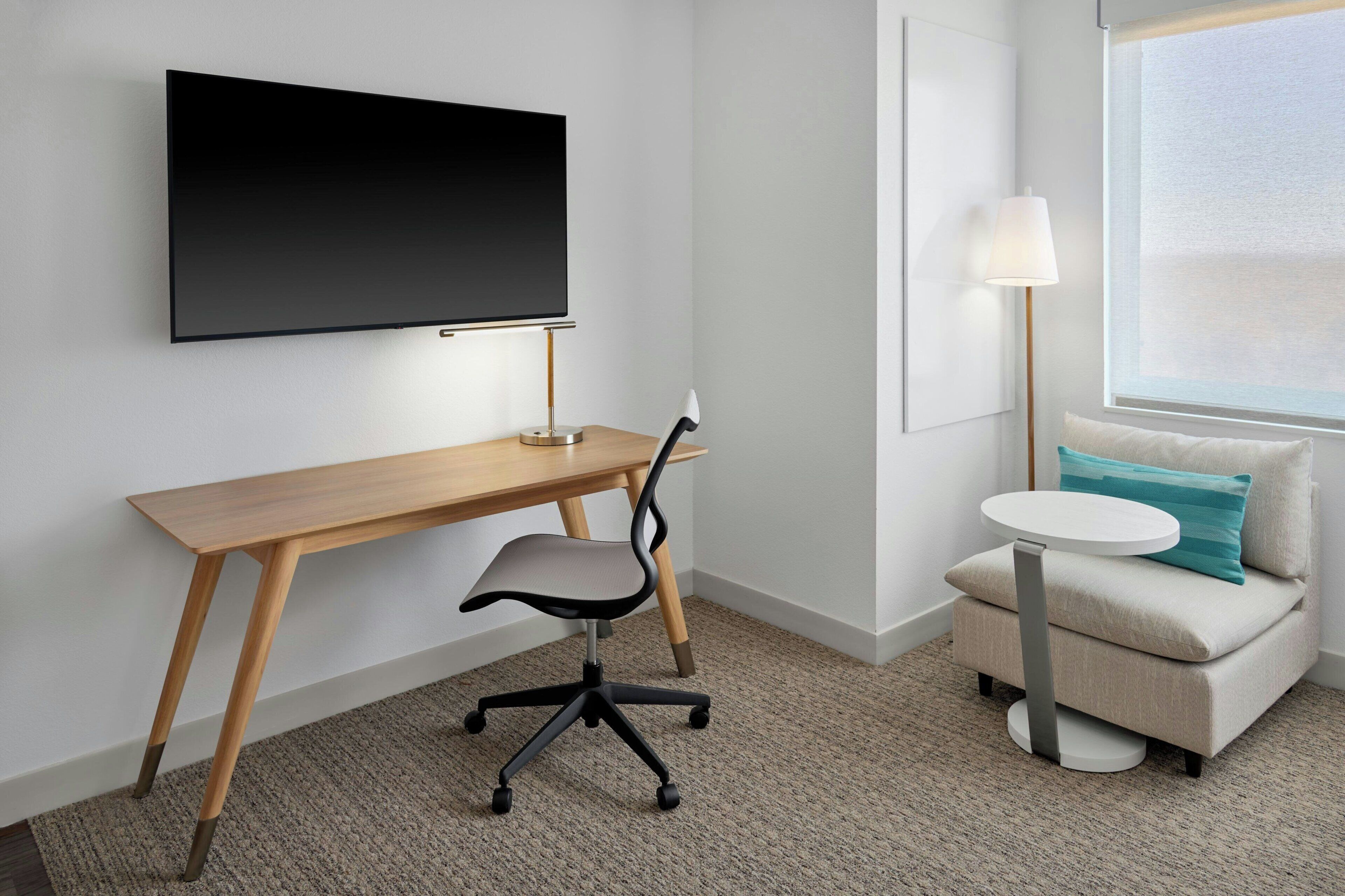 Une chambre d’hôtel avec un bureau en bois, une chaise moderne, un téléviseur à écran plat fixé au mur, une petite table ronde, une lampe et un canapé avec un coussin.