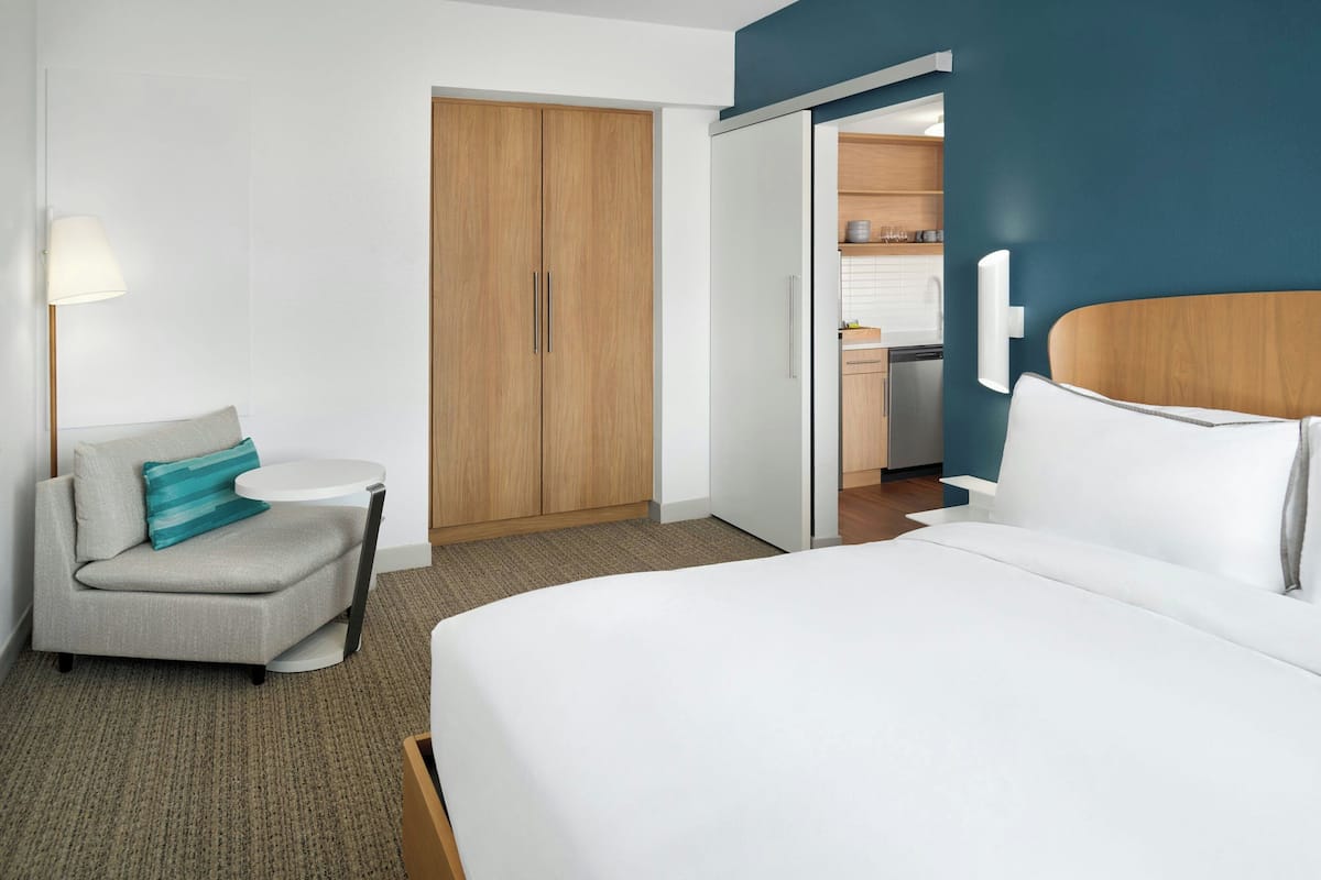 A hotel room with a bed, a sofa, a desk, a wardrobe, and a kitchenette in the background.
