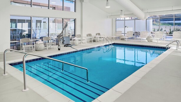 An indoor swimming pool with clear blue water, surrounded by white lounge chairs and a sunbed.