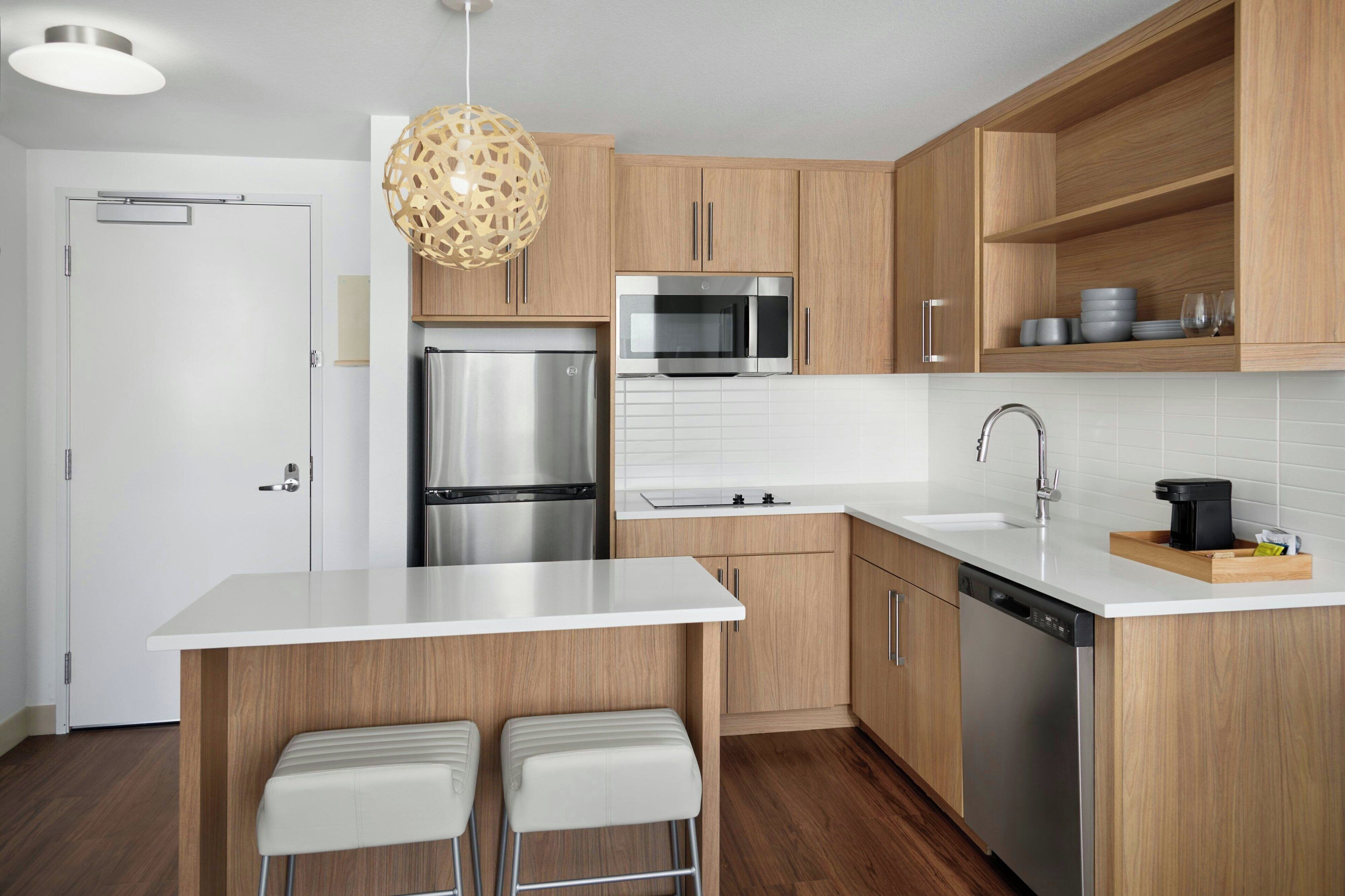 A modern kitchen with wooden cabinets, stainless steel appliances, a white countertop, and two bar stools.