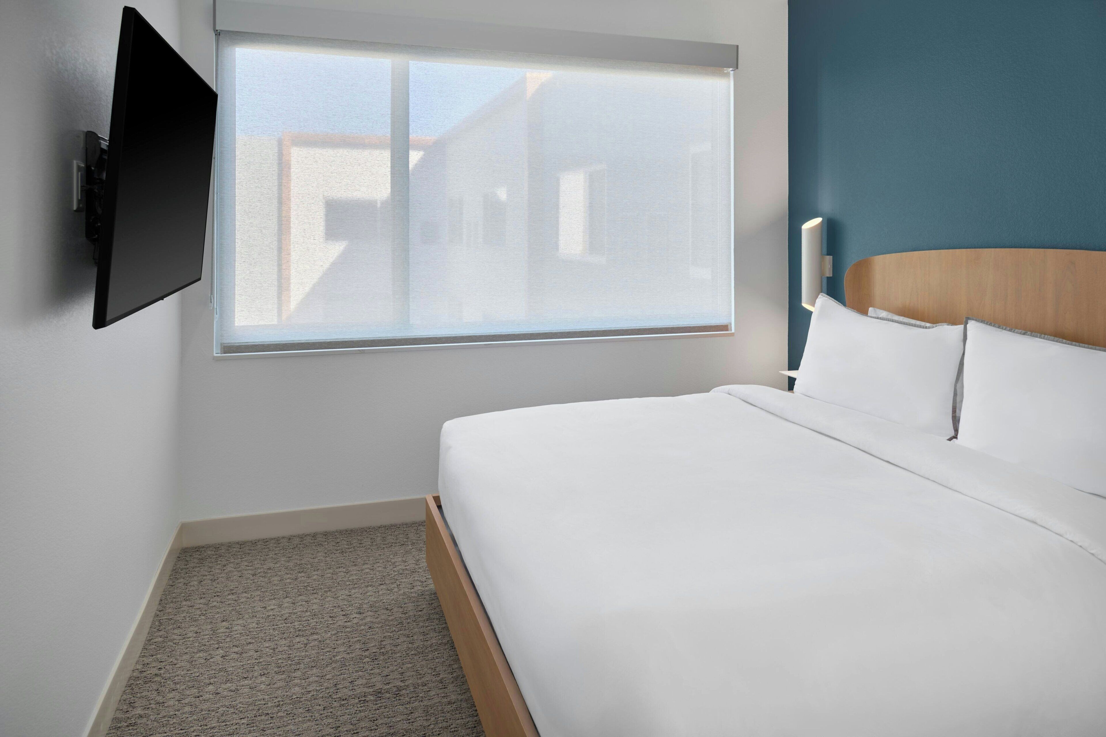 A hotel room with a bed, a TV mounted on the wall, and a window with a view of buildings.
