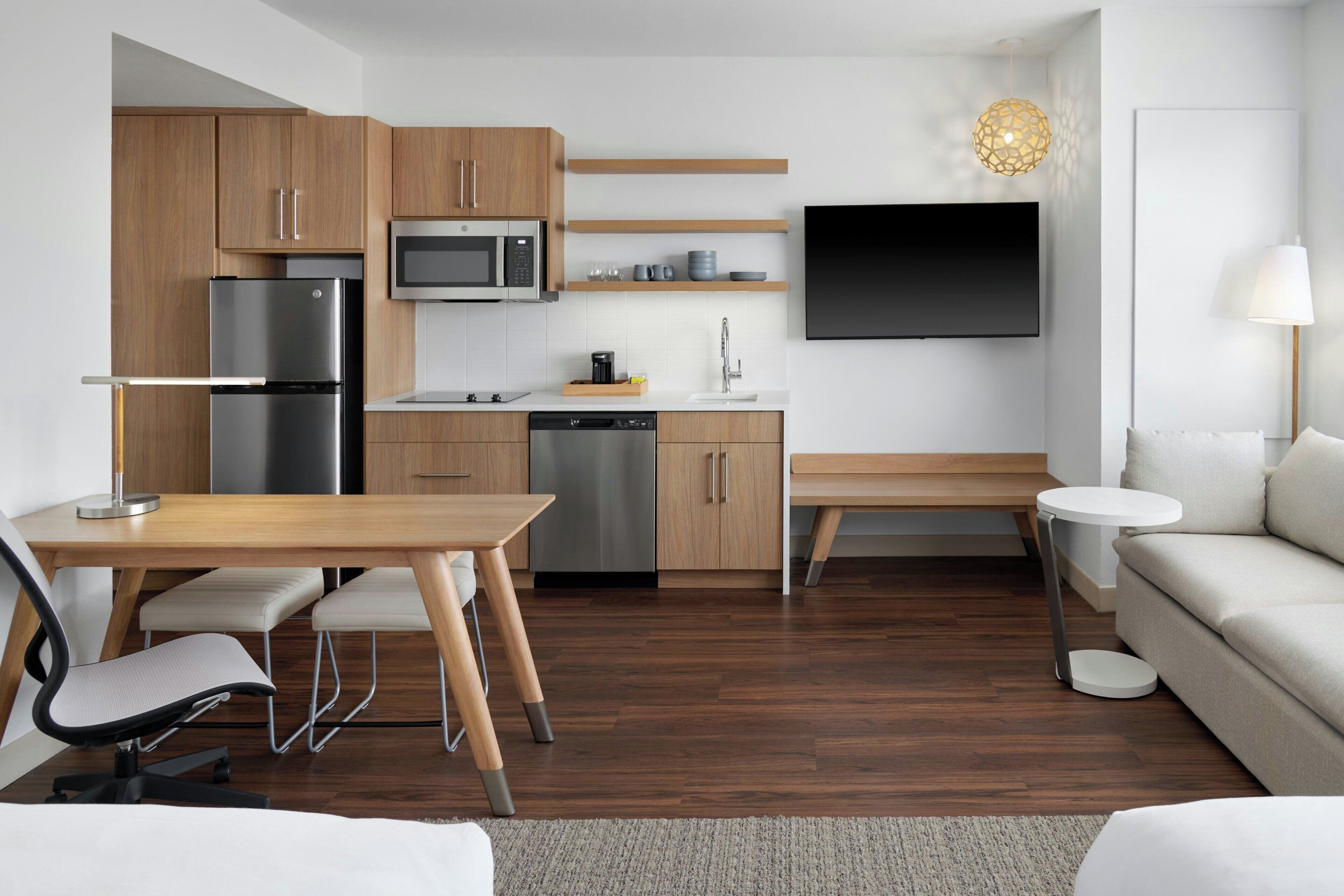 A modern kitchen with wooden cabinets, a stainless steel refrigerator, a microwave, and a dishwasher. There is a dining table with chairs, a bench, and a round white side table. A flat-screen TV is mounted on the wall above the kitchen counter.