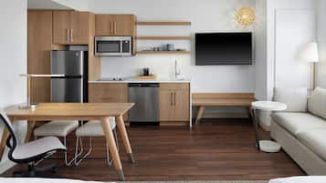 Eine moderne Küche mit Holzschränken, einem Edelstahlkühlschrank, einer Mikrowelle und einem Geschirrspüler. Ein Esstisch mit Stühlen, eine Bank und ein runder weißer Beistelltisch sind ebenfalls vorhanden. Ein Flachbildfernseher ist über der Küchenarbeitsfläche an der Wand montiert.