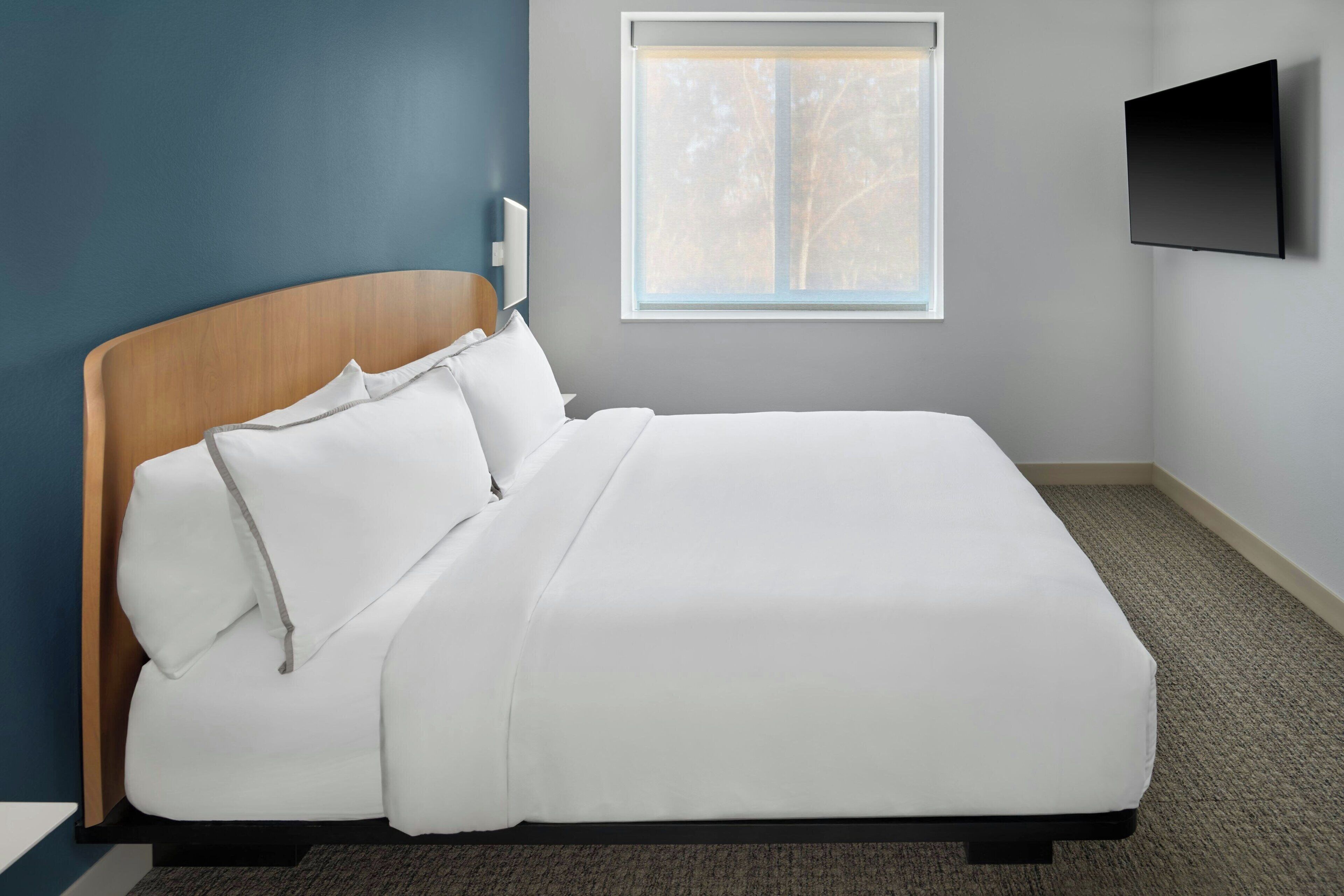 A neatly made bed with white bedding in a room with a blue accent wall, a window, and a flat-screen TV mounted on the wall.