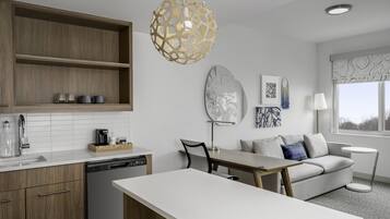 A modern kitchen with wooden cabinets, a white countertop, and a dishwasher. There is a dining area with a table and chairs, a sofa, and a lamp.