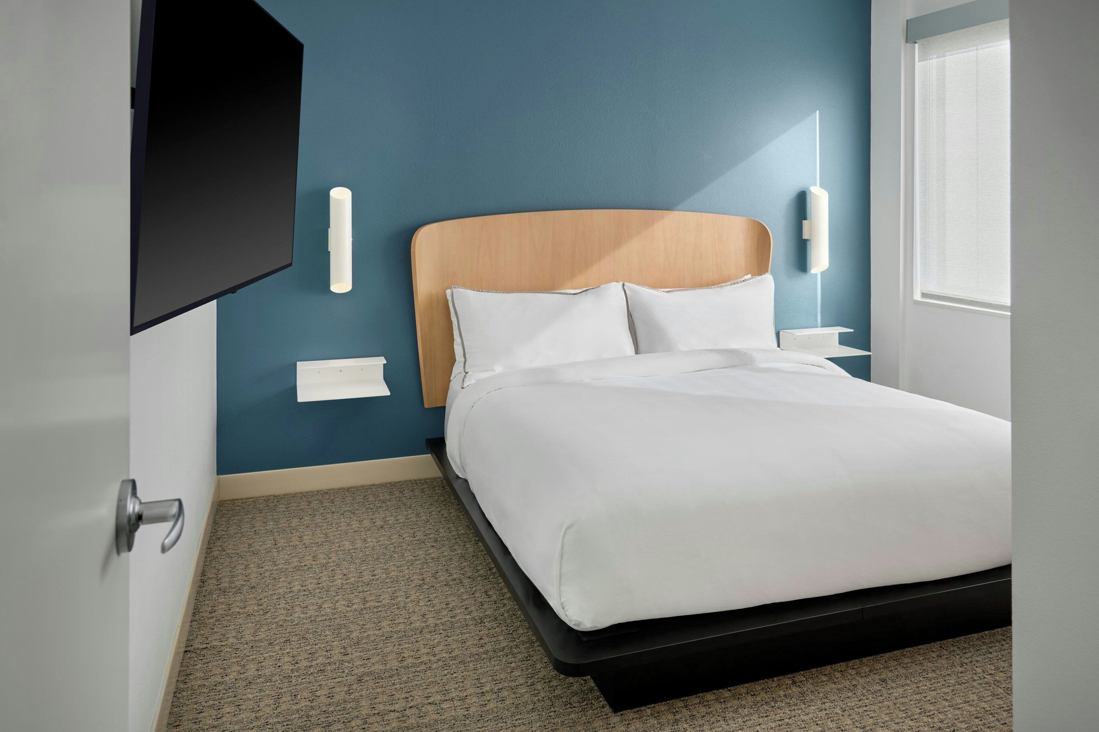 Ein modernes Hotelzimmer mit Bett, Flachbildfernseher an der Wand, Nachttisch mit Lampe und einem Fenster mit Jalousien.