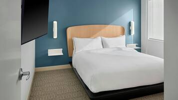 Ein modernes Hotelzimmer mit Bett, Flachbildfernseher an der Wand, Nachttisch mit Lampe und einem Fenster mit Jalousien.