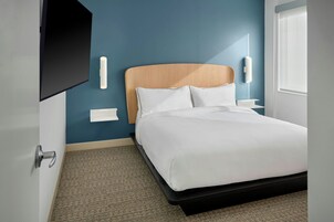 Ein modernes Hotelzimmer mit Bett, Flachbildfernseher an der Wand, Nachttisch mit Lampe und einem Fenster mit Jalousien.