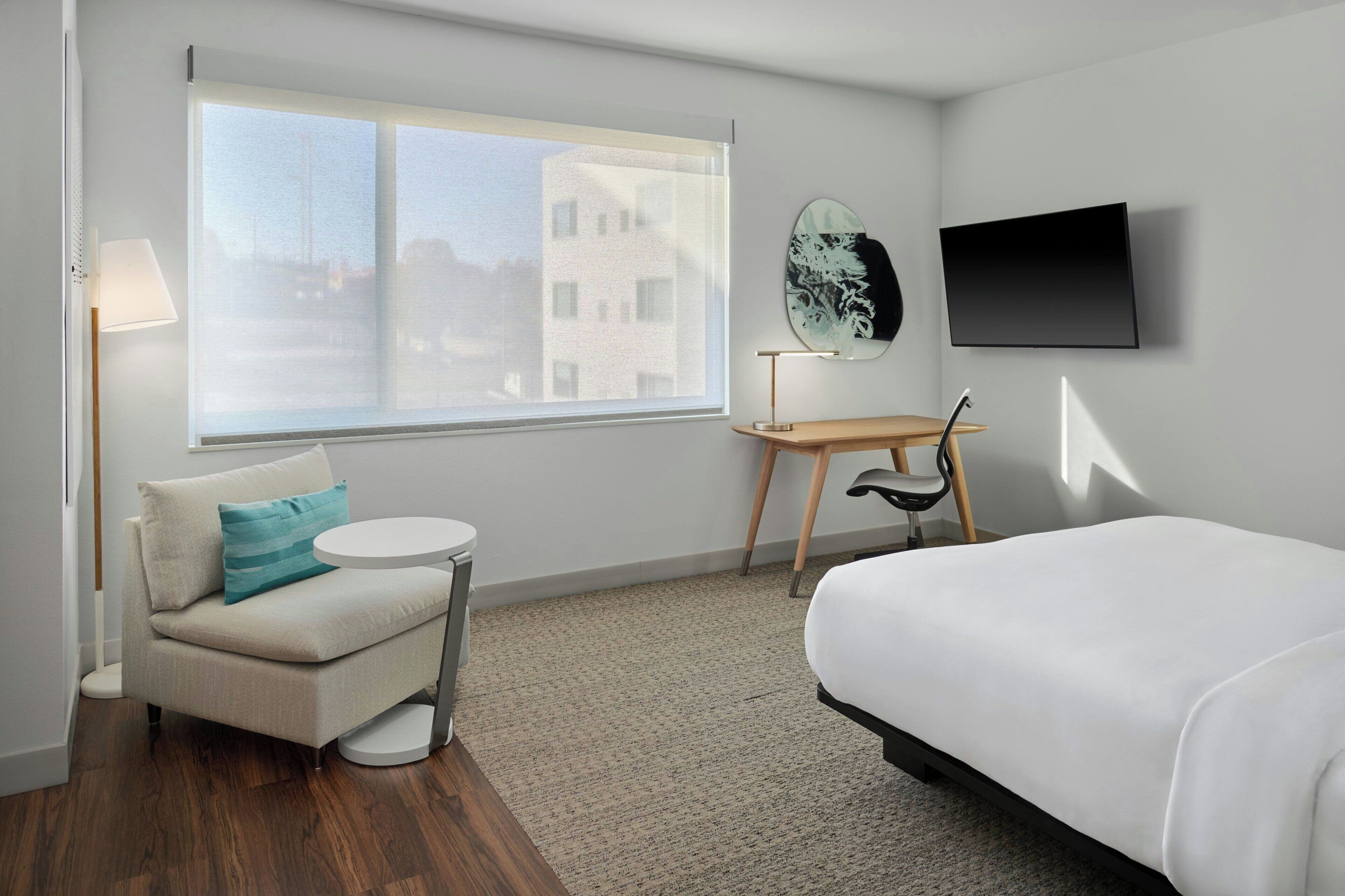 Habitación de hotel con cama, sofá, una mesita redonda, una silla, televisor y un amplio ventanal con vistas a edificios.