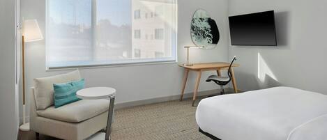 Habitación de hotel con cama, sofá, una mesita redonda, una silla, televisor y un amplio ventanal con vistas a edificios.