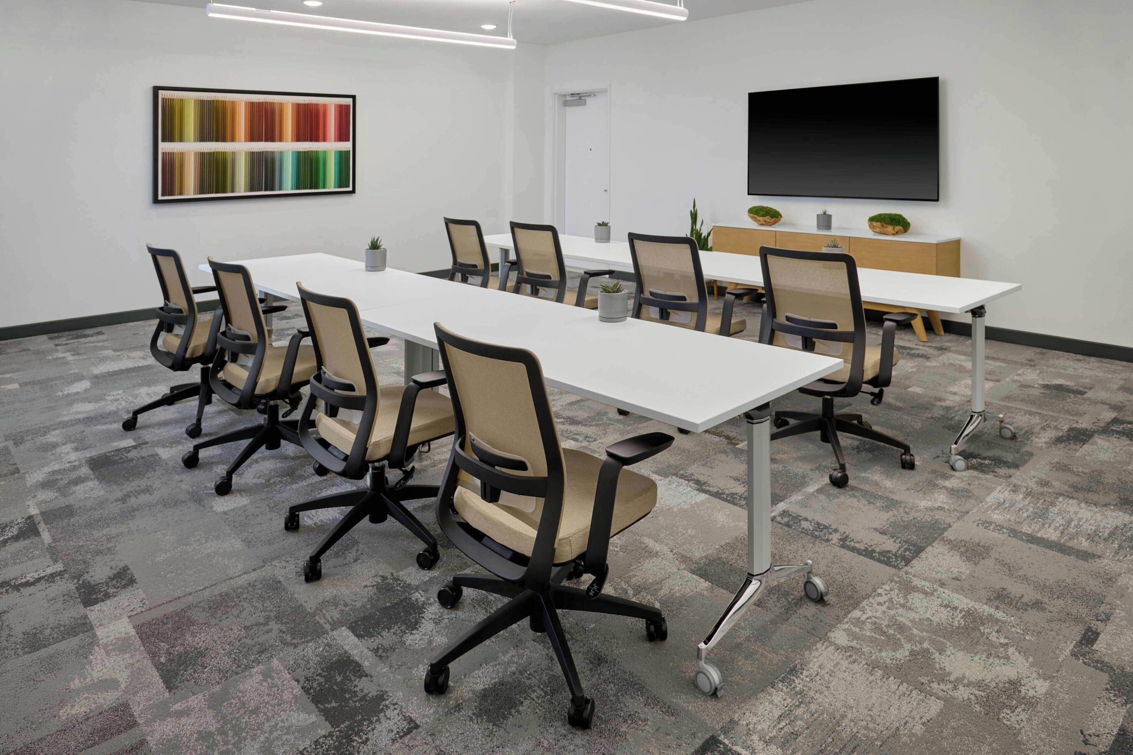 A conference room with a long table, multiple chairs, a TV screen, and a colorful abstract painting on the wall.