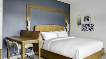 A modern hotel room with a wooden bed frame, white bedding, a wooden desk with two chairs, and a large mirror above the bed.