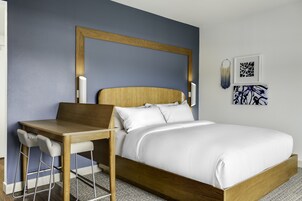 A modern hotel room with a wooden bed frame, white bedding, a wooden desk with two chairs, and a large mirror above the bed.