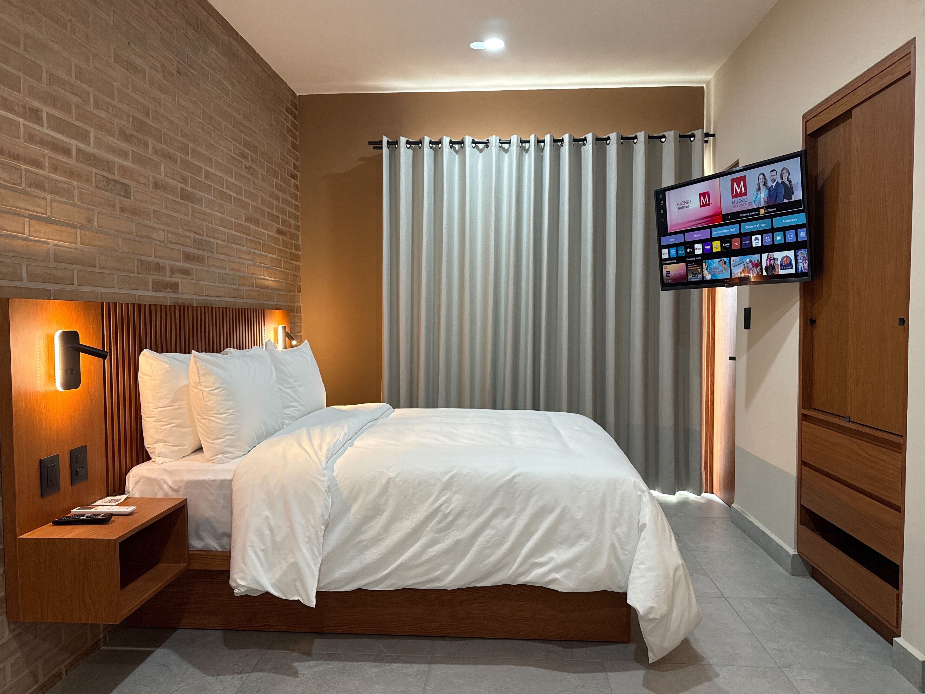 A hotel room with a brick accent wall, a bed with white bedding, a nightstand with a remote control, a television mounted on the wall displaying a menu, and a wooden wardrobe.