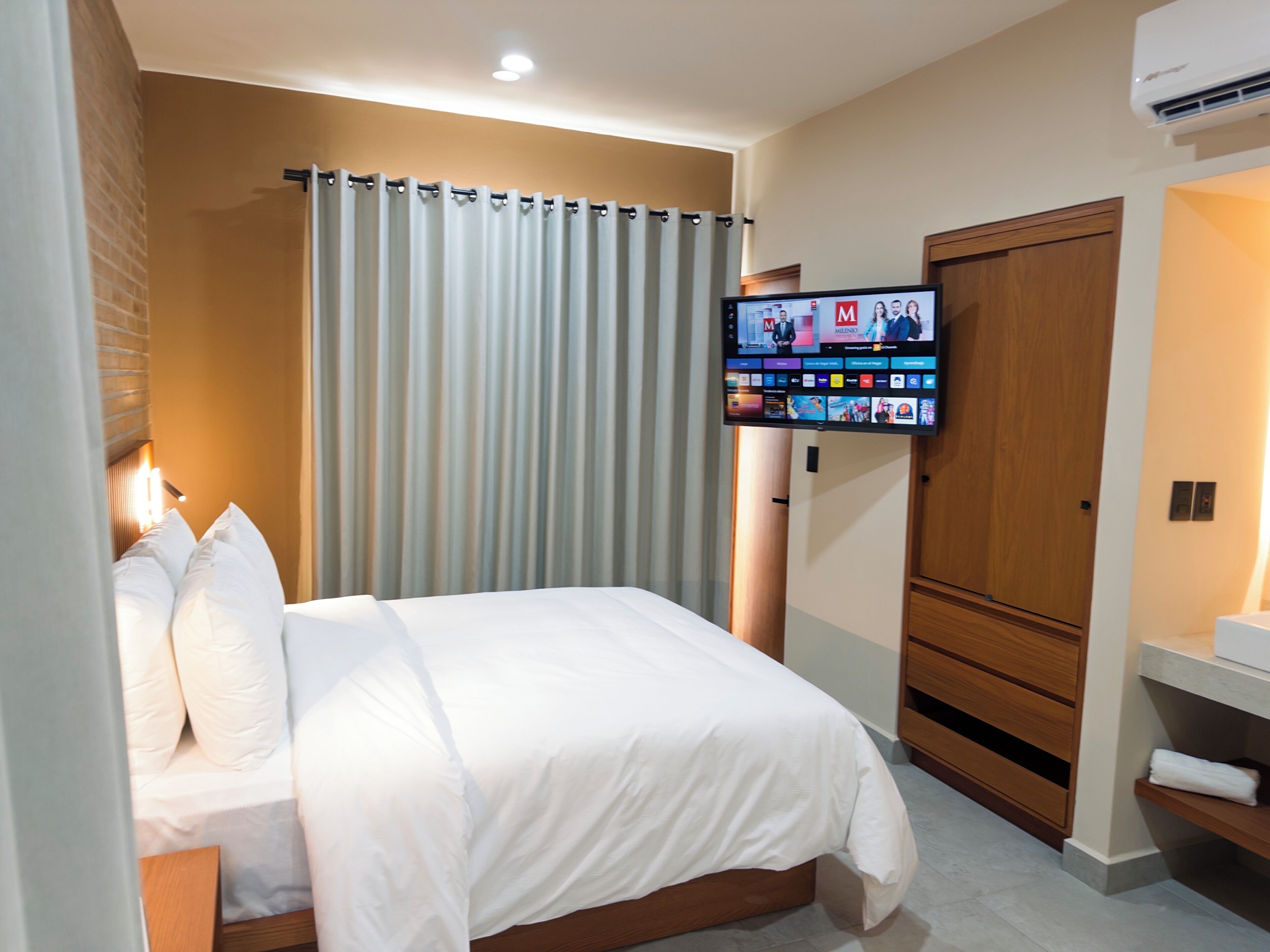 A hotel room with a bed, a television mounted on the wall, a wooden wardrobe, and a bathroom with a sink and towel rack.