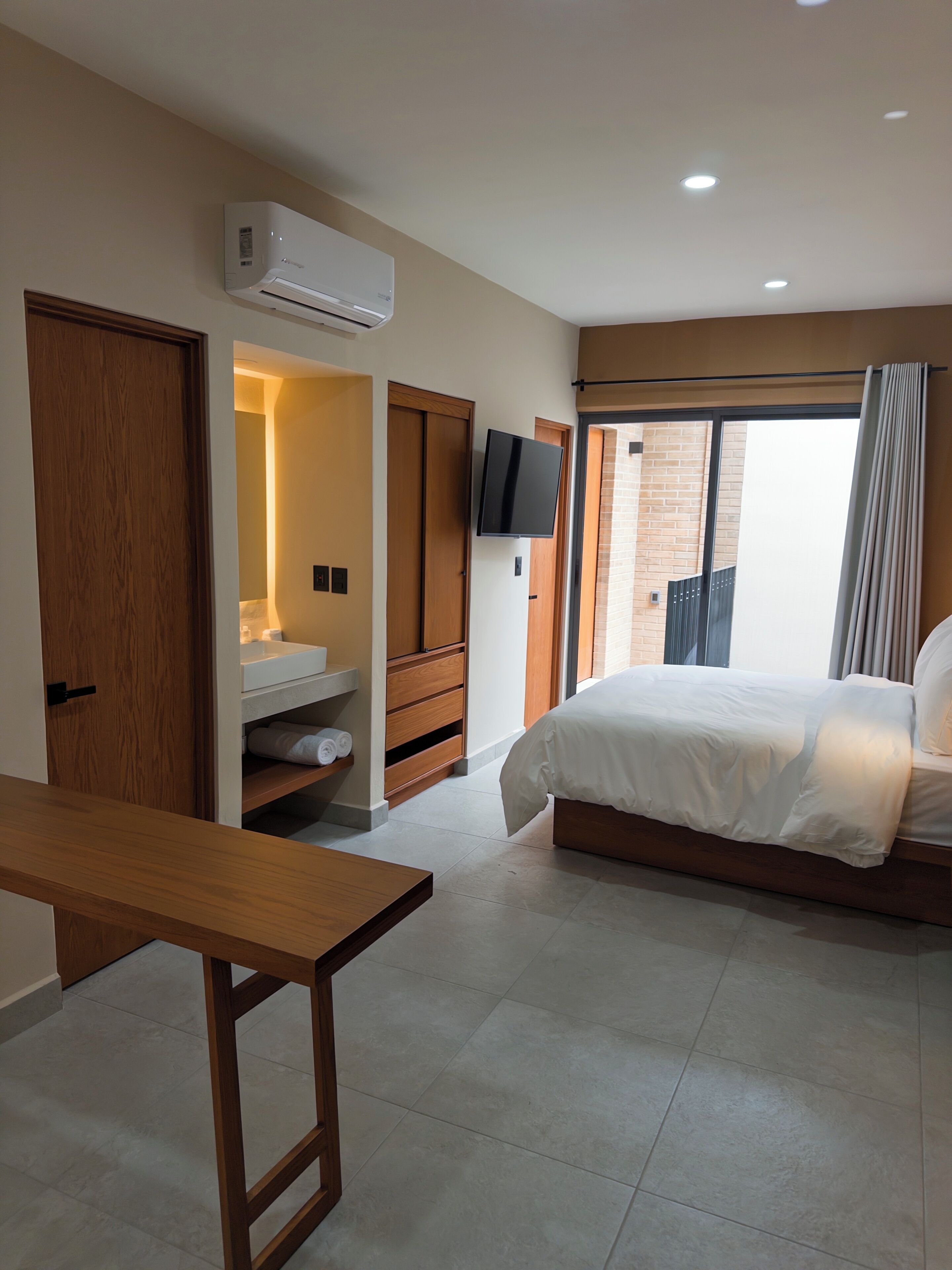 Une chambre d’hôtel moderne équipée d’un lit, d’une télévision, d’une salle de bain avec un lavabo et un miroir, et d’une porte en bois.