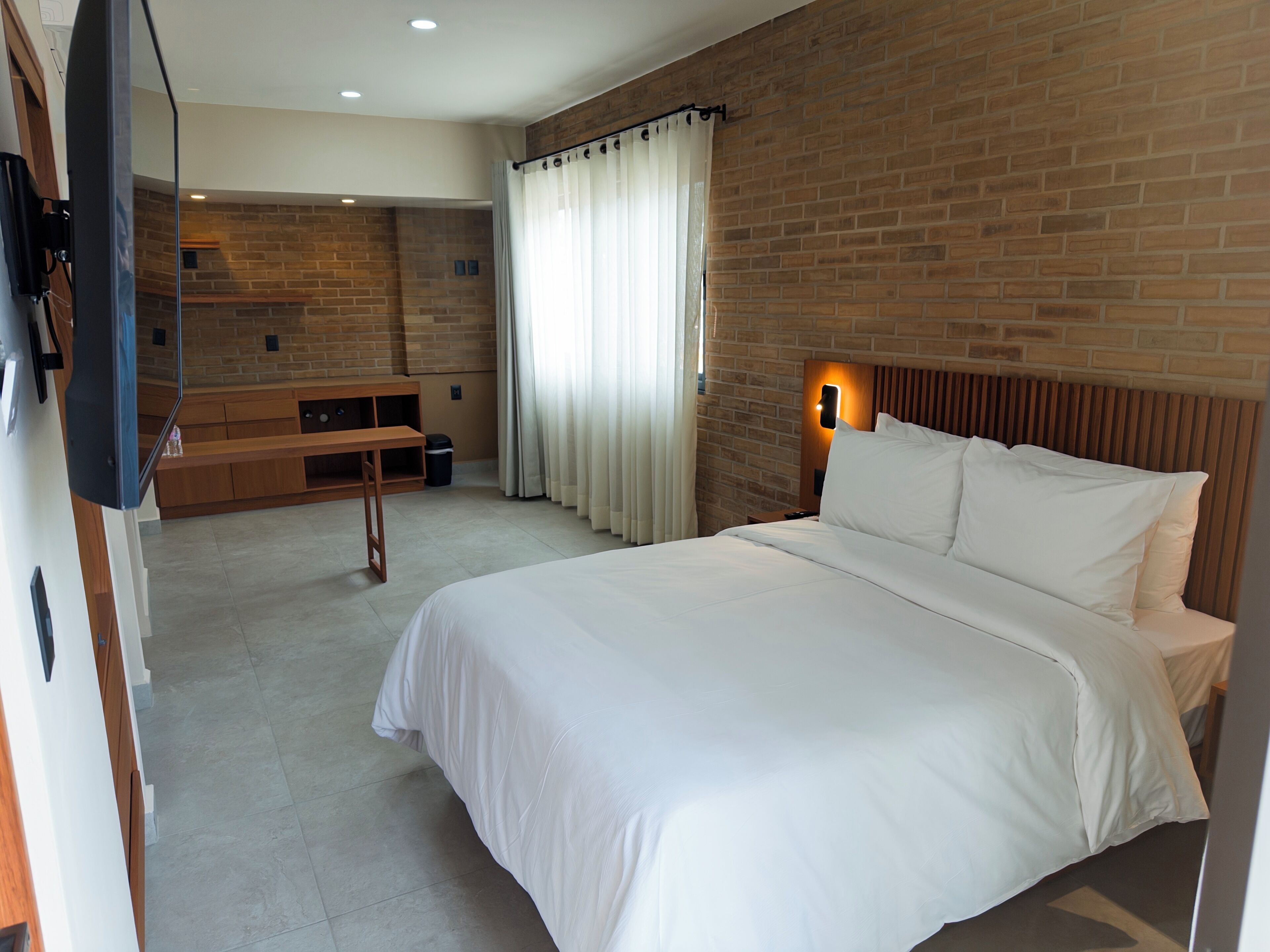 Une chambre d’hôtel avec un mur en briques, un lit avec une literie blanche, un téléviseur fixé au mur, un banc en bois et une fenêtre avec des rideaux.